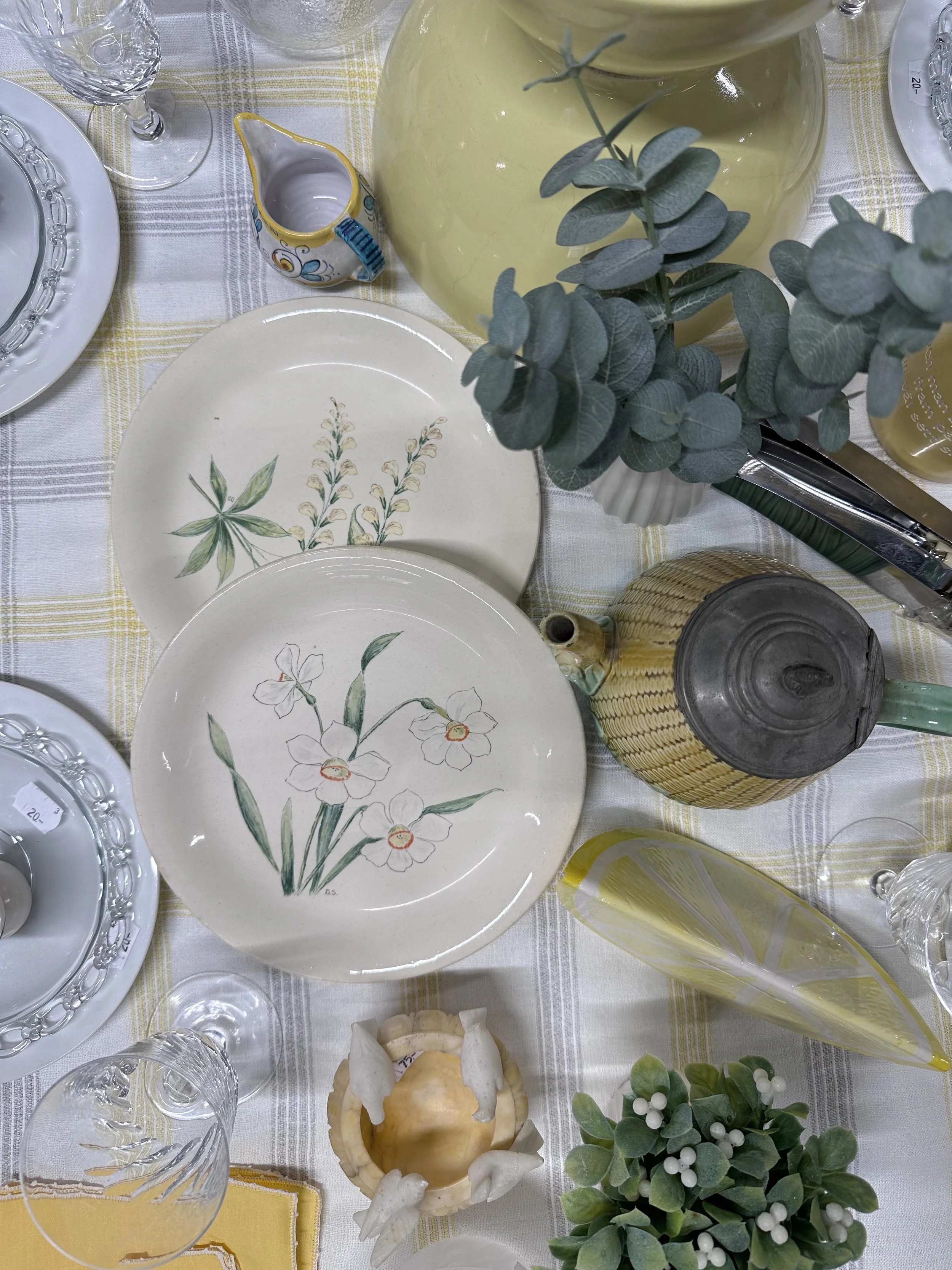 Porslinsfat med blomster- och bladmönster, gröna växter, olika glas och en liten vas på ett bord med ljusgult och vit rutigt duk.