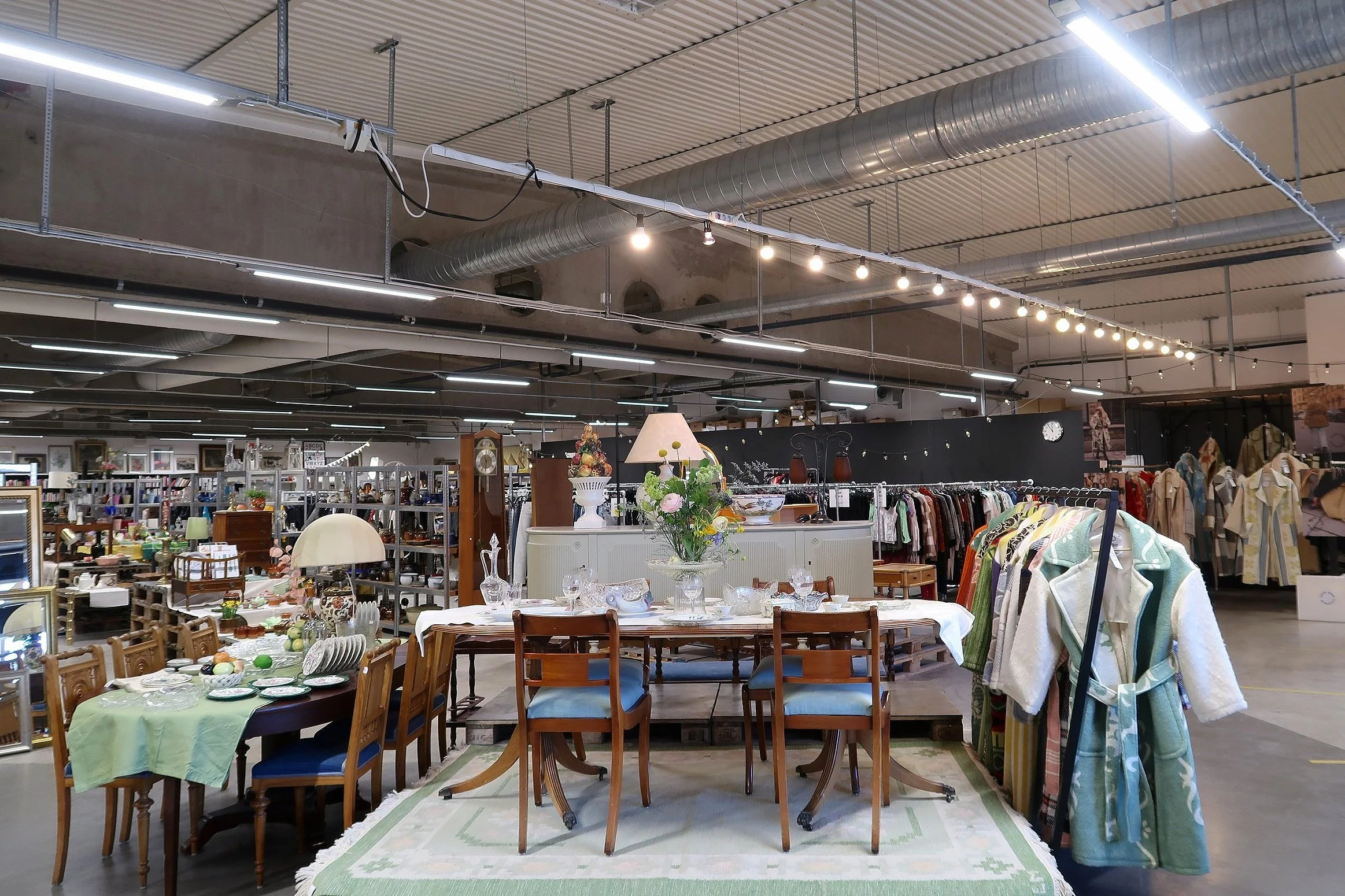 Inomhusbild av ett second hand- eller vintagevaruhus med möbler, kläder och heminredning, inklusive ett bord med porslin och blommor, och en klädhängare med badrockar.