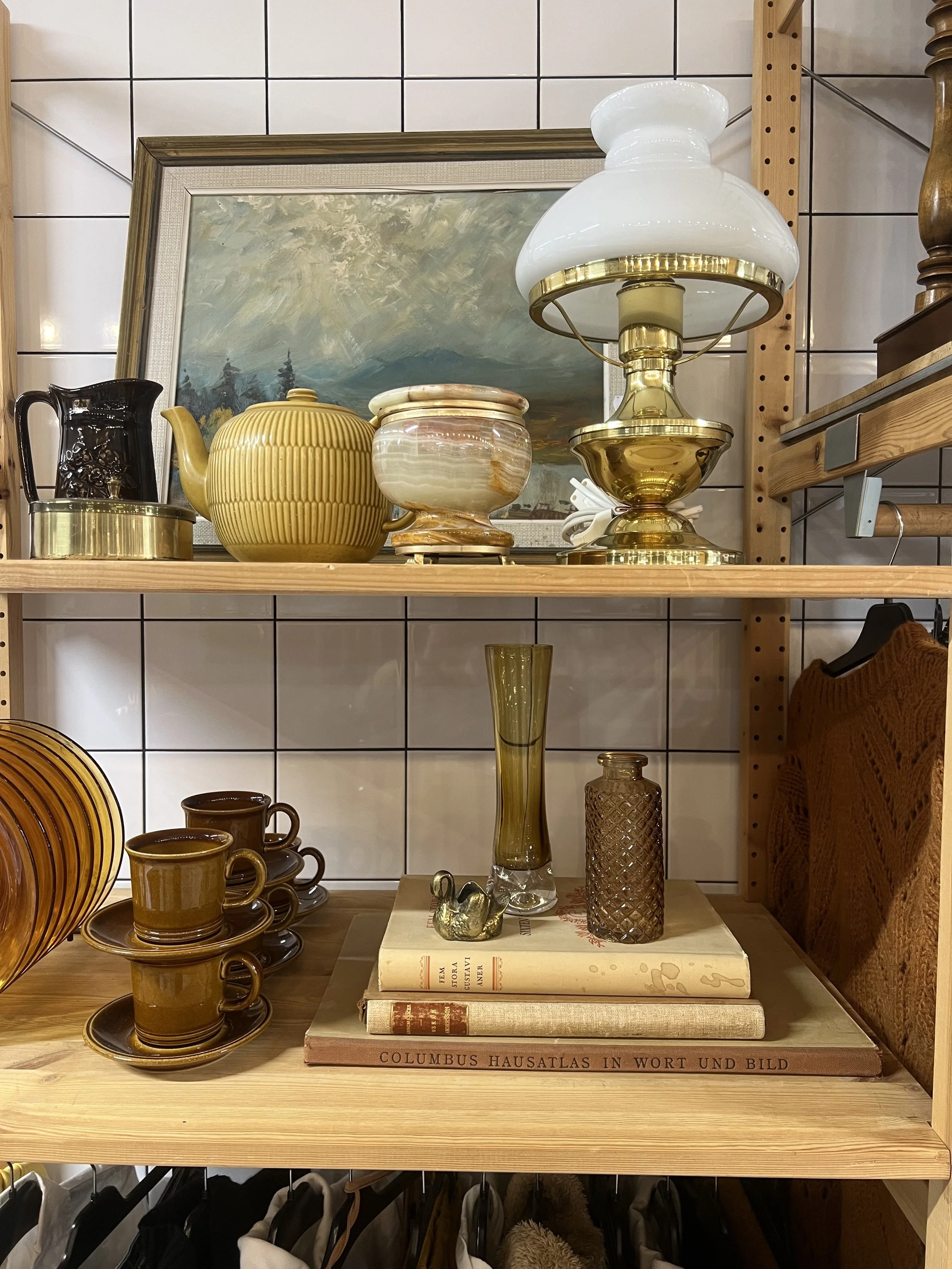 Vägg med vitklinkers och tre hyllor med dekorativa föremål, inklusive ett målat landskapsfoto, på den övre hyllan finns keramik och en glasskål, på den nedre hyllan finns glasvaser, böcker, en skulptur och en samling koppar med underkoppar.