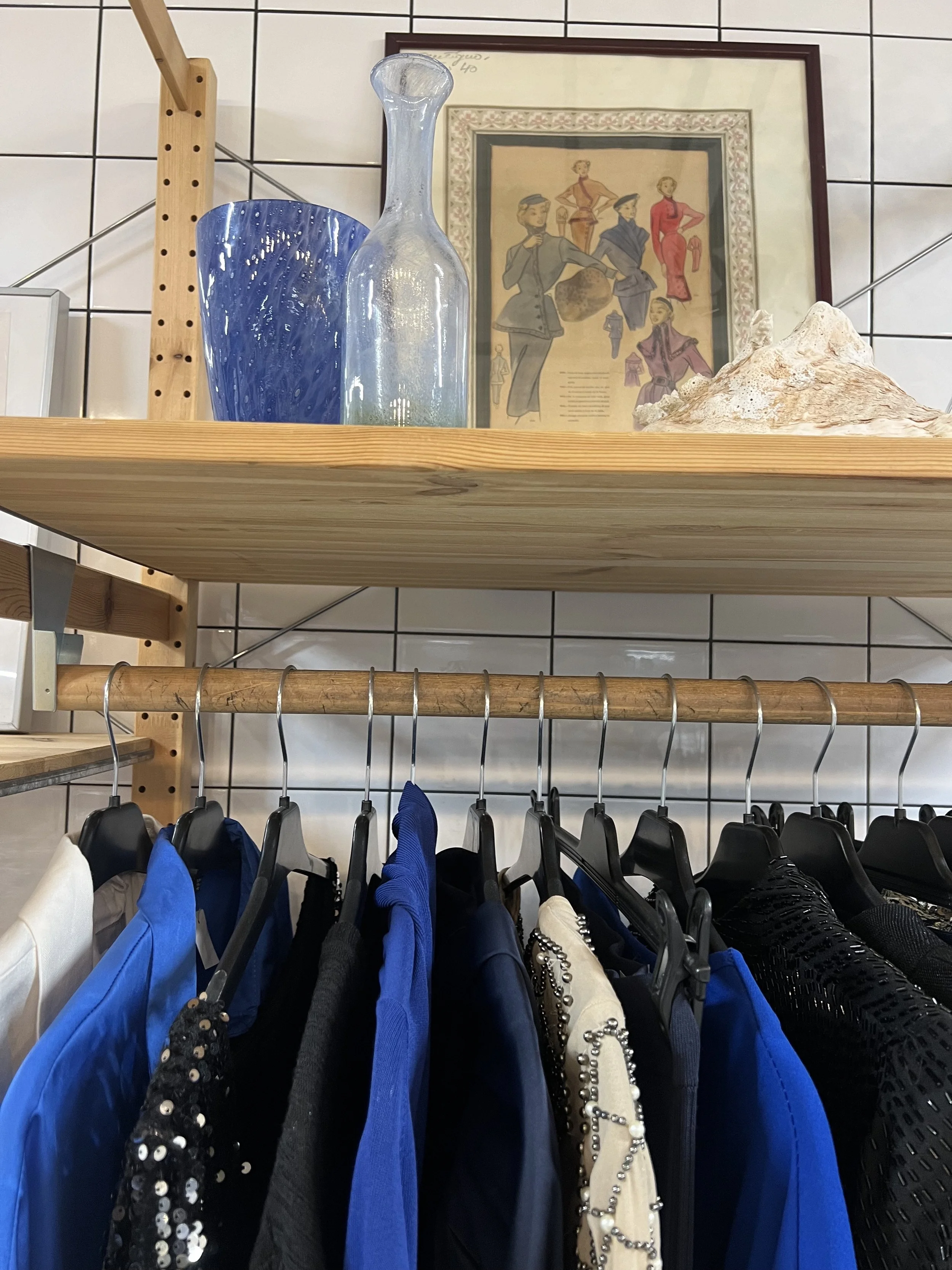 Skjortor i olika färger och material hängande på en klädstång, med dekorativa glasflaskor och konst på vägg i bakgrunden.