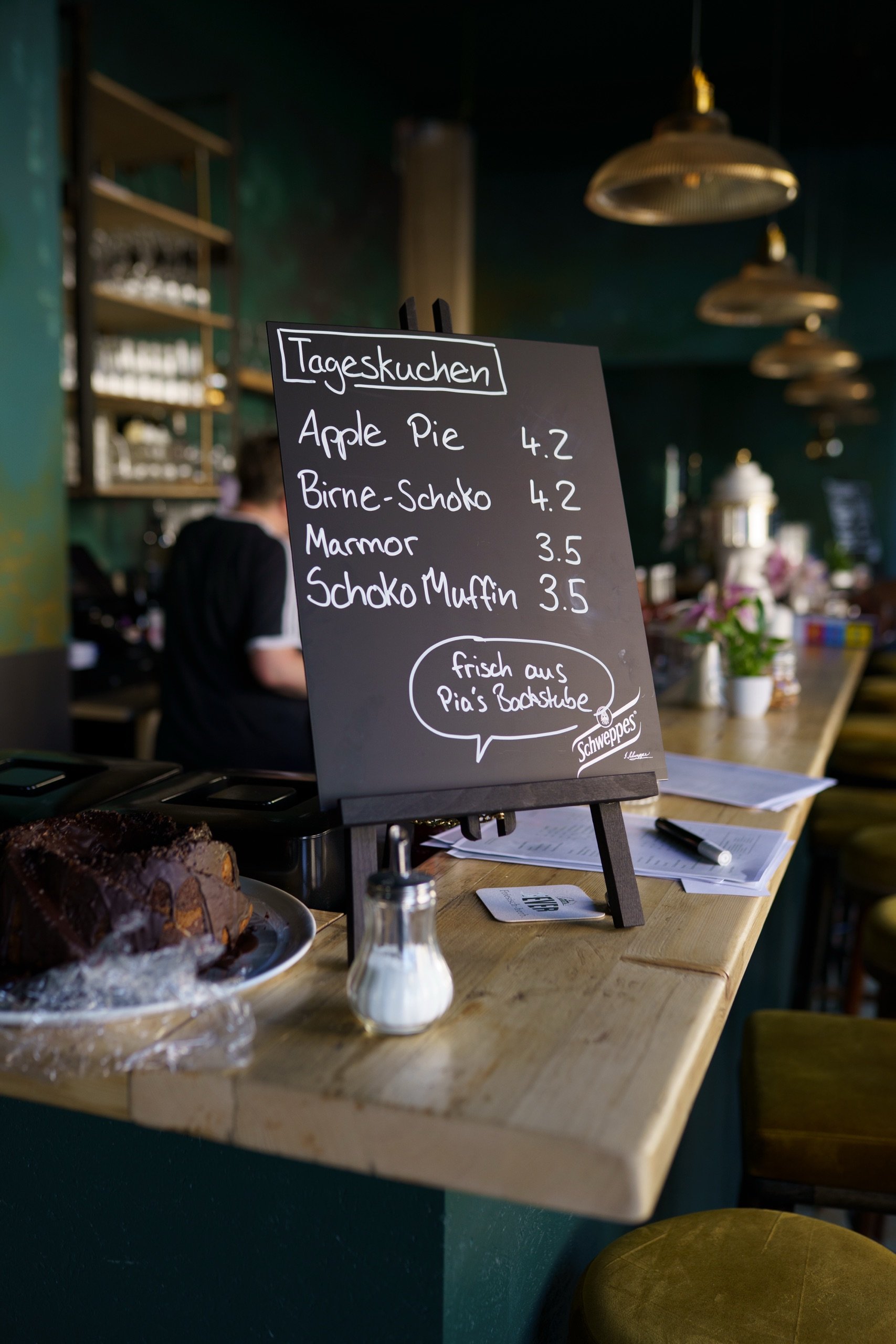 Schwarzes Menübrett mit "Tageskuchen" Angeboten neben einem Schokoladenkuchen auf Tresen in Café.