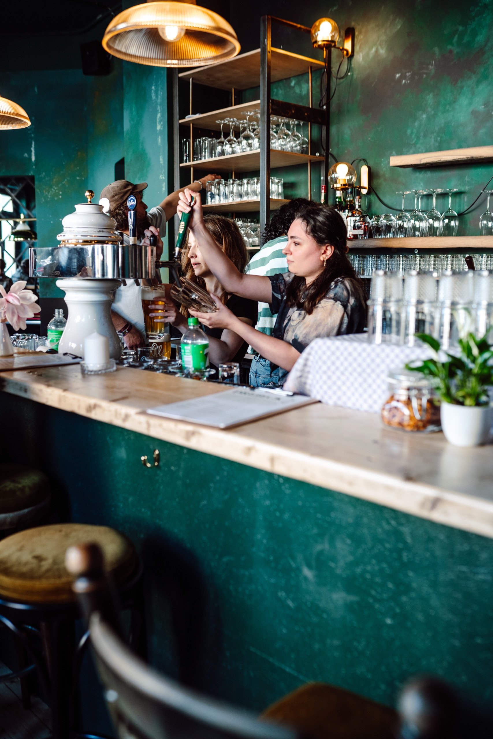 Zwei Frauen arbeiten hinter einer Bar, zapfen Bier und bedienen Gäste. Im Hintergrund sind Gläser und Flaschen auf Regalen. Die Bar ist in warmen, grünen Farben dekoriert.