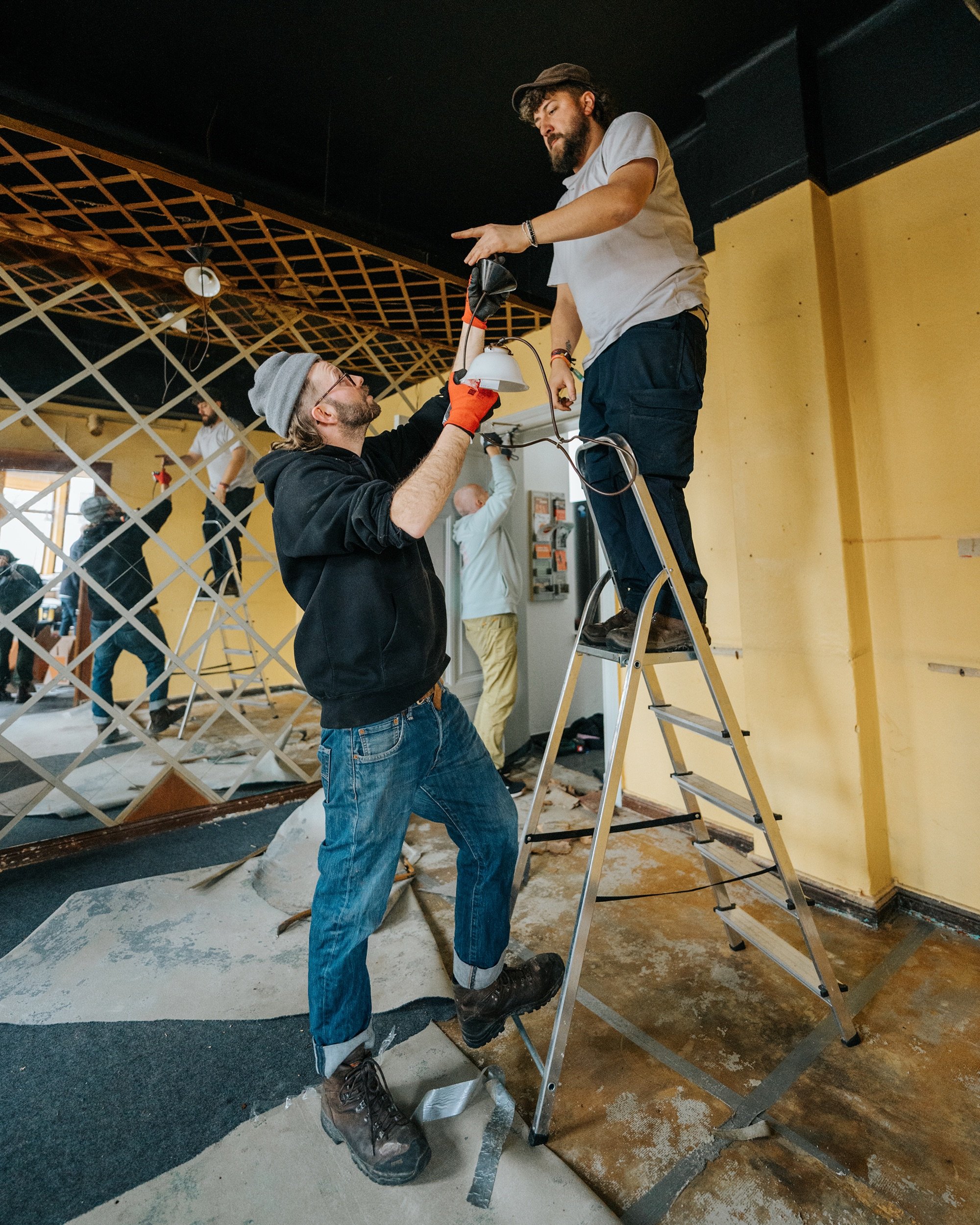 Zwei Männer arbeiten an Renovierungsprojekt, einer steht auf einer Leiter und der andere reicht ihm ein Werkzeug, baustellenähnliches Umfeld.