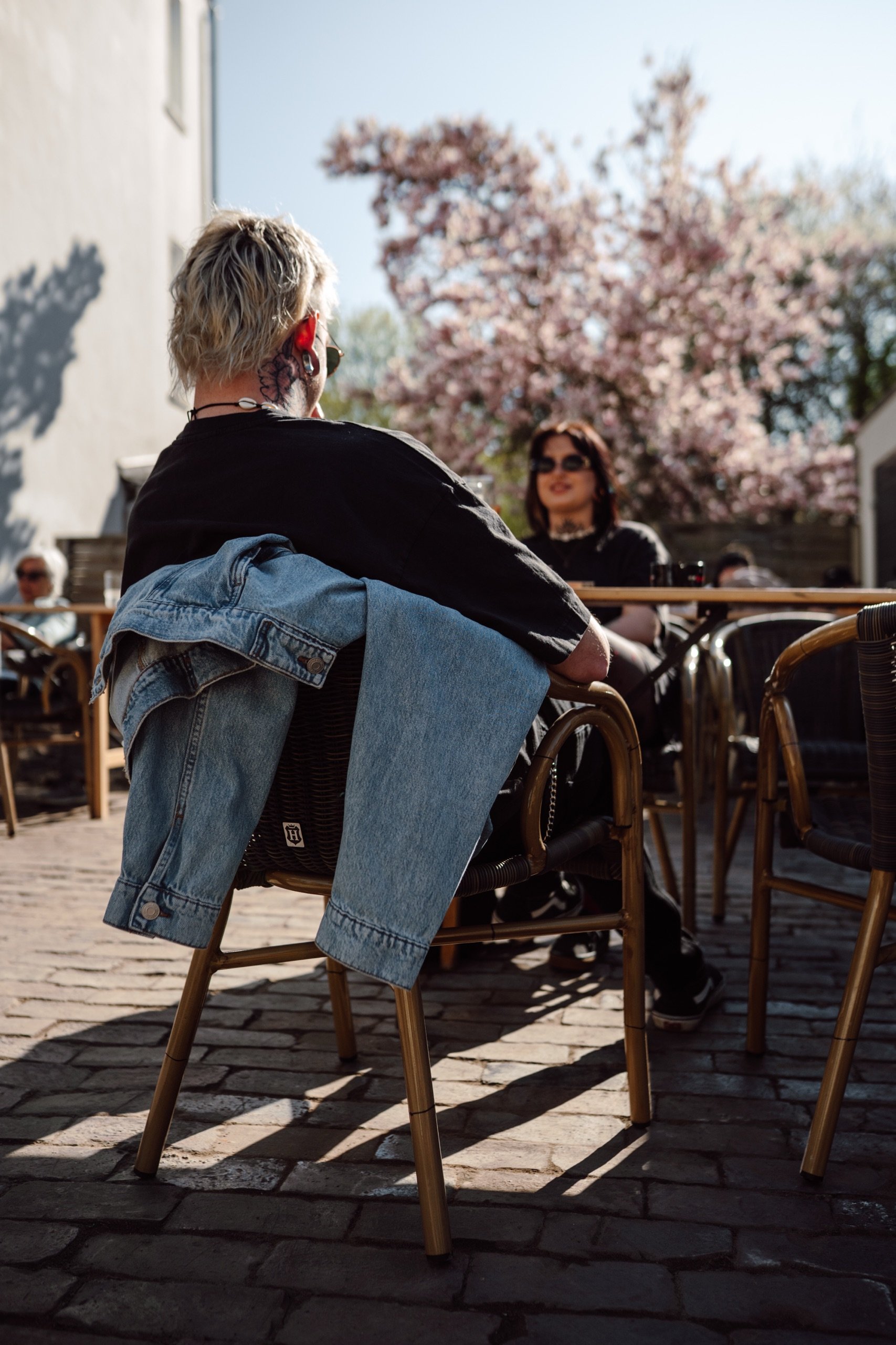 Personen sitzen in einem Café im Freien. Im Vordergrund sitzt jemand mit einer Jeansjacke über dem Stuhl, im Hintergrund blühender Baum und sonniges Wetter.