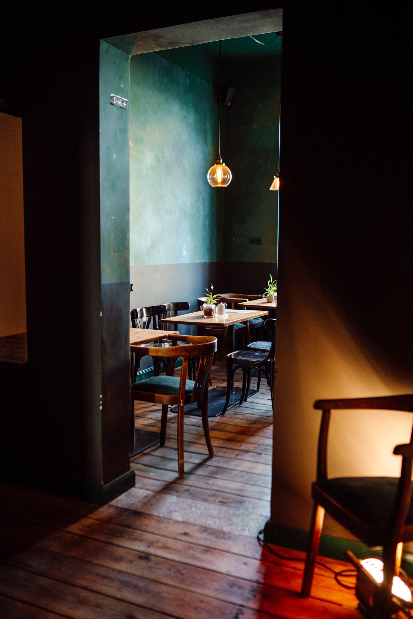 Ein gemütlicher Innenraum eines Cafés oder Restaurants mit Holzboden, Holztischen und Stühlen. Der Raum ist schwach beleuchtet, mit hängenden Lampen und grünen Wänden. Pflanzen stehen auf den Tischen zur Dekoration.