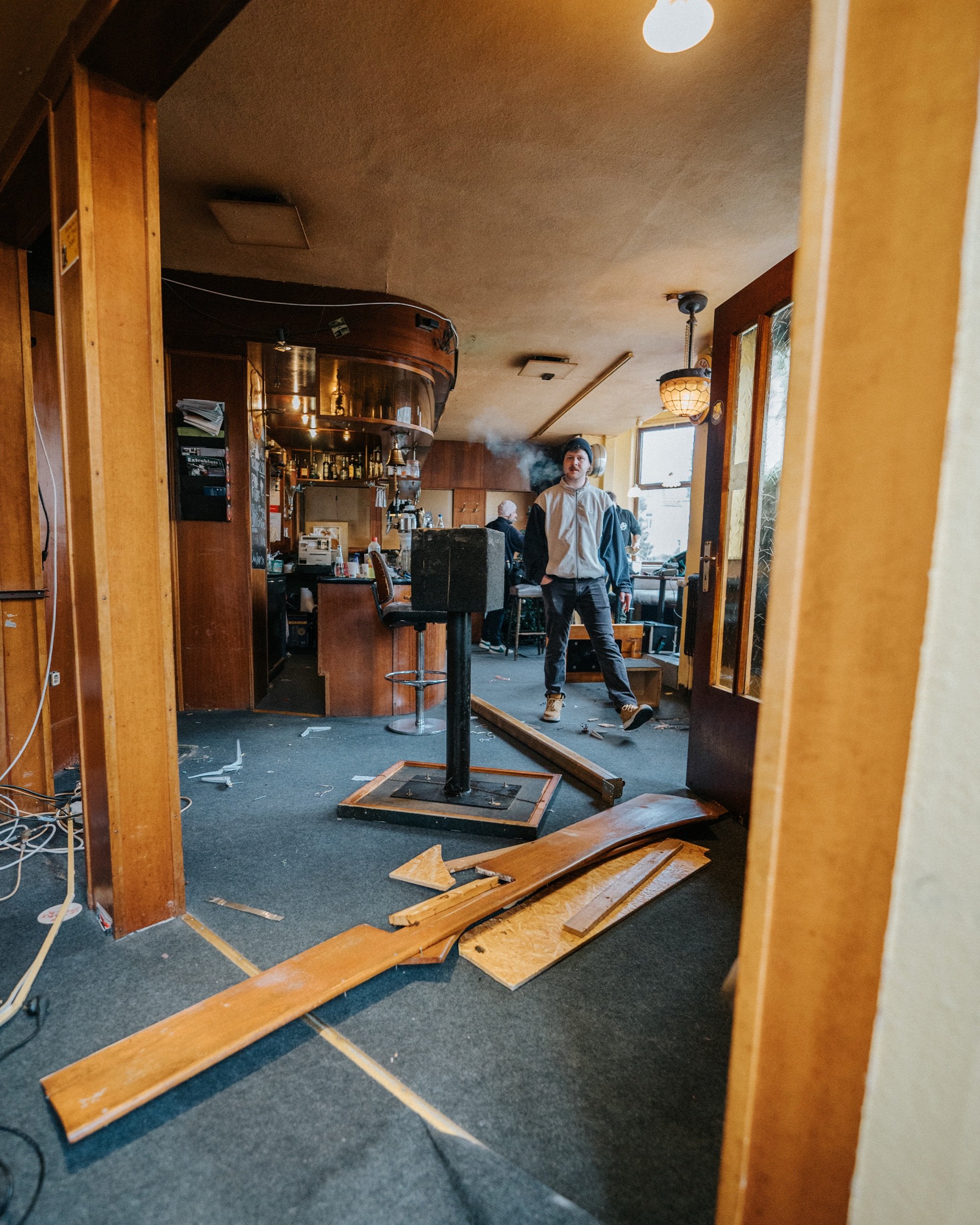 Innenraum einer Bar mit Holzmöbeln, beschädigten Böden und Holzstücken auf dem Boden, eine Person steht rauchend in der Nähe der Bar.