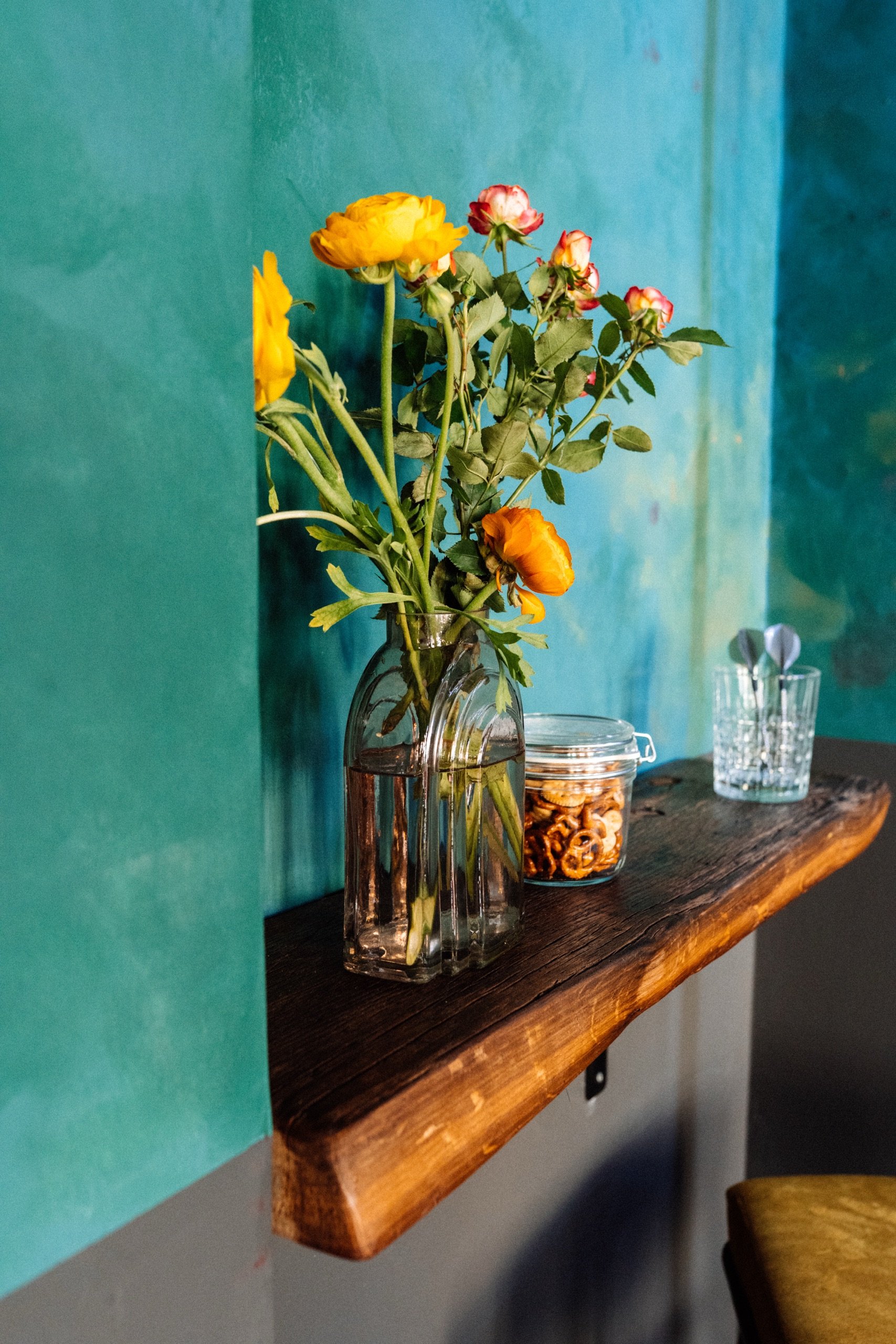 Ein Glasvase mit gelben und rosa Blumen auf einem hölzernen Regal vor einer türkisblauen Wand. Daneben steht ein Glas mit Brezeln und ein Glas mit Besteck.