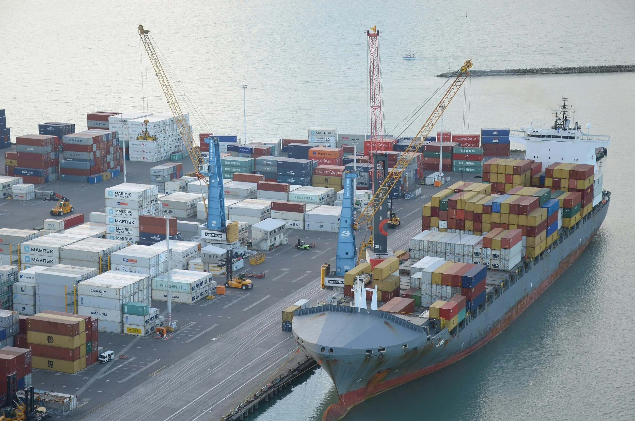 Large cargo ship docked at port with numerous stacked shipping containers and cranes for loading and unloading, adjacent to a body of water.