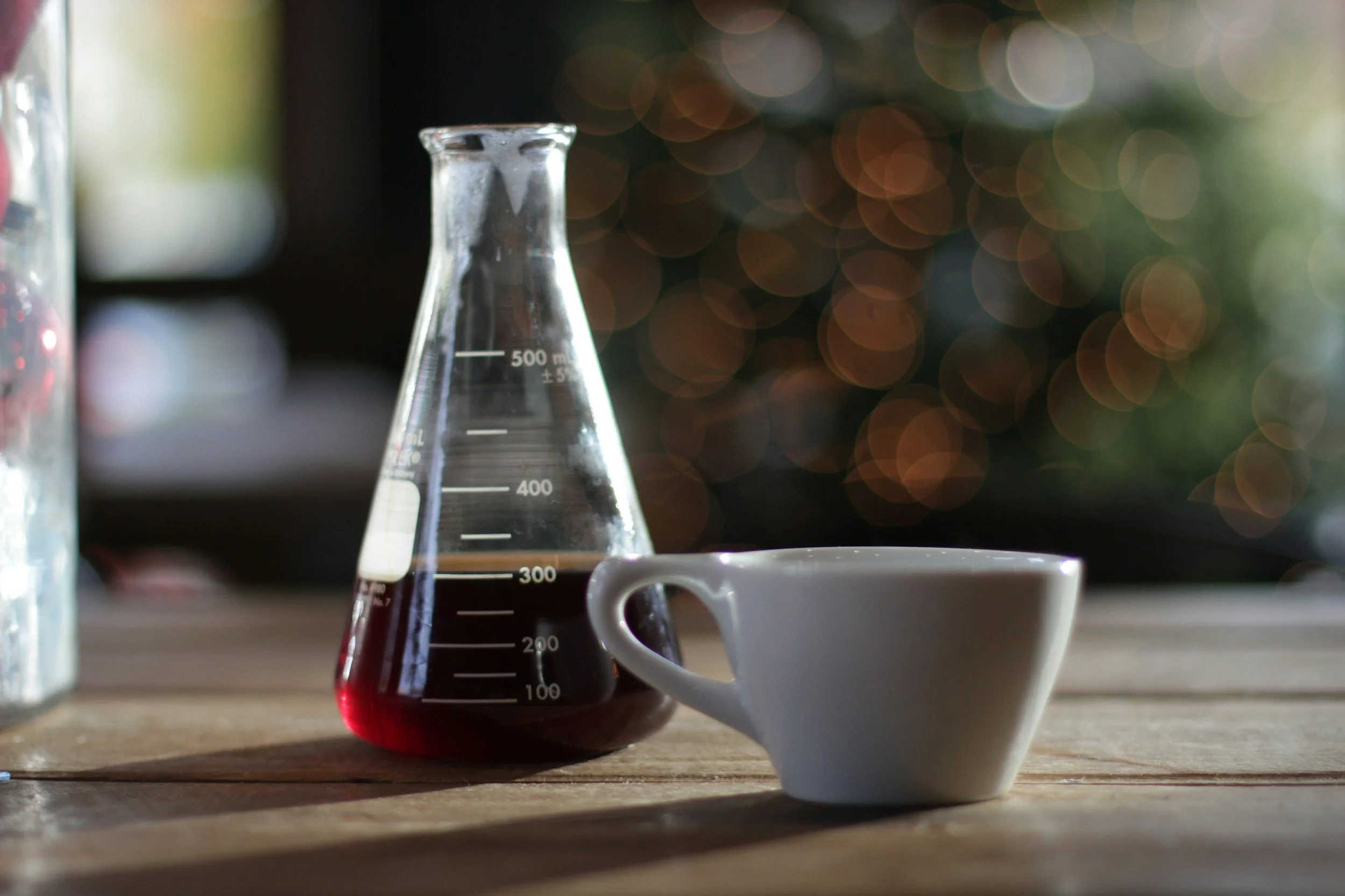 A beaker with dark brown liquid and a white coffee cup on a wooden table with blurred festive lights in the background.