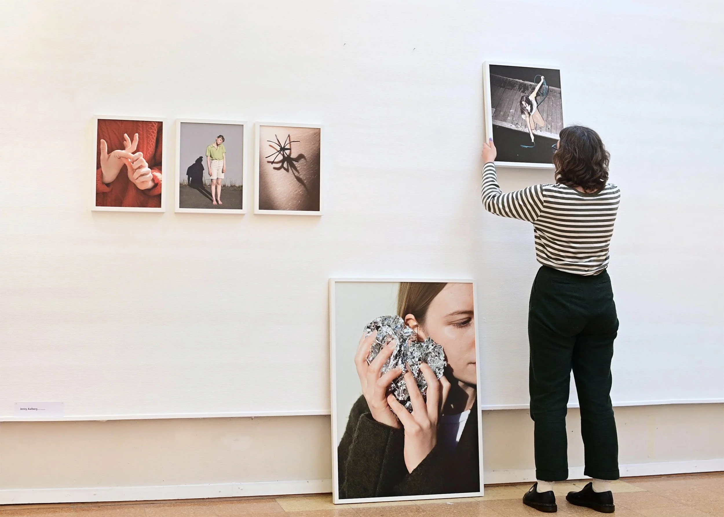 En person henger opp et bilde i et galleri. Det er flere bilder på veggen og et stort fotografi står på gulvet.