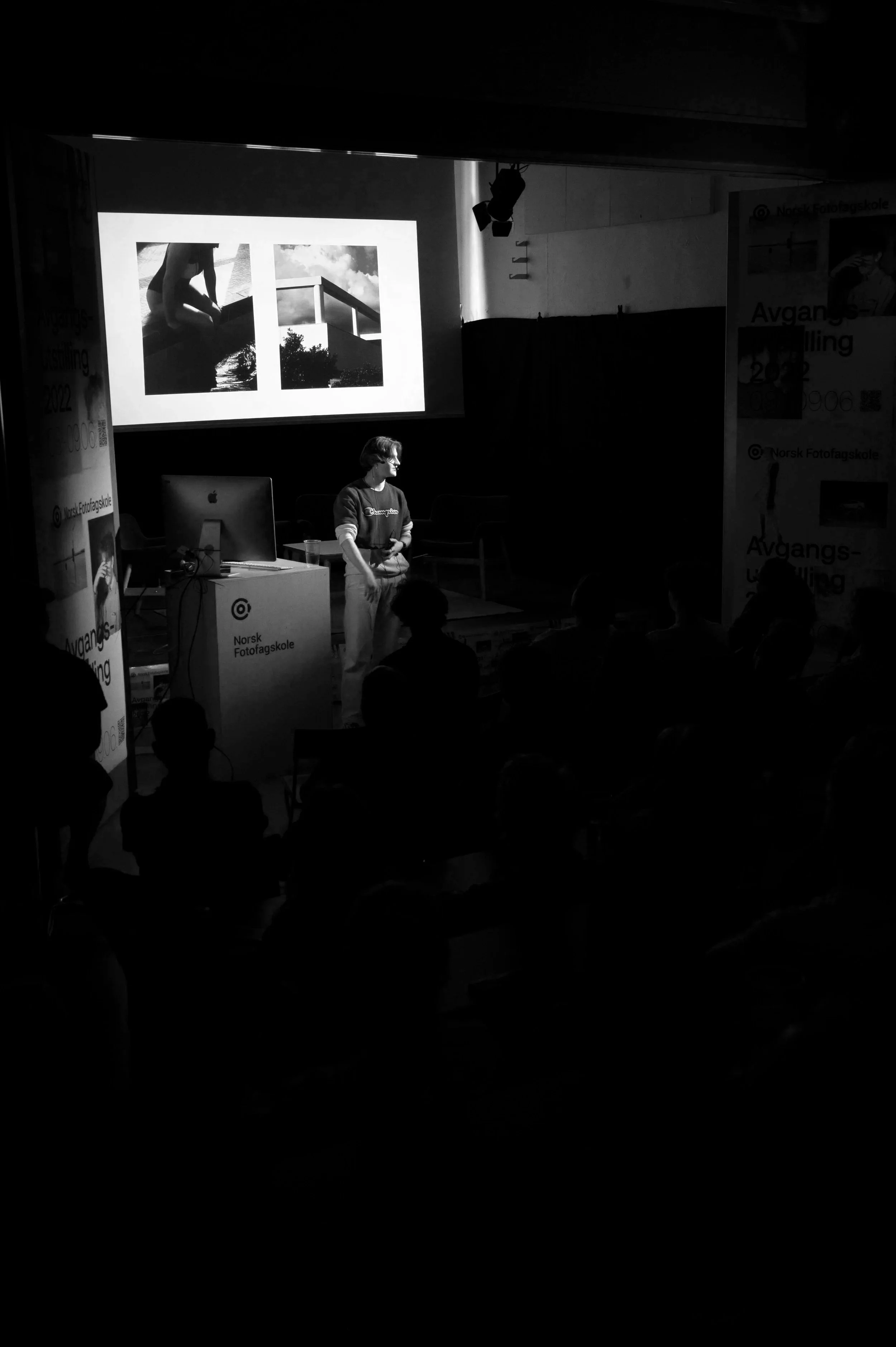 En person holder en presentasjon foran en skjerm med bilder av en person som sitter ved vann, og en struktur med trapp og himmel. Publikum ses i bakgrunnen.