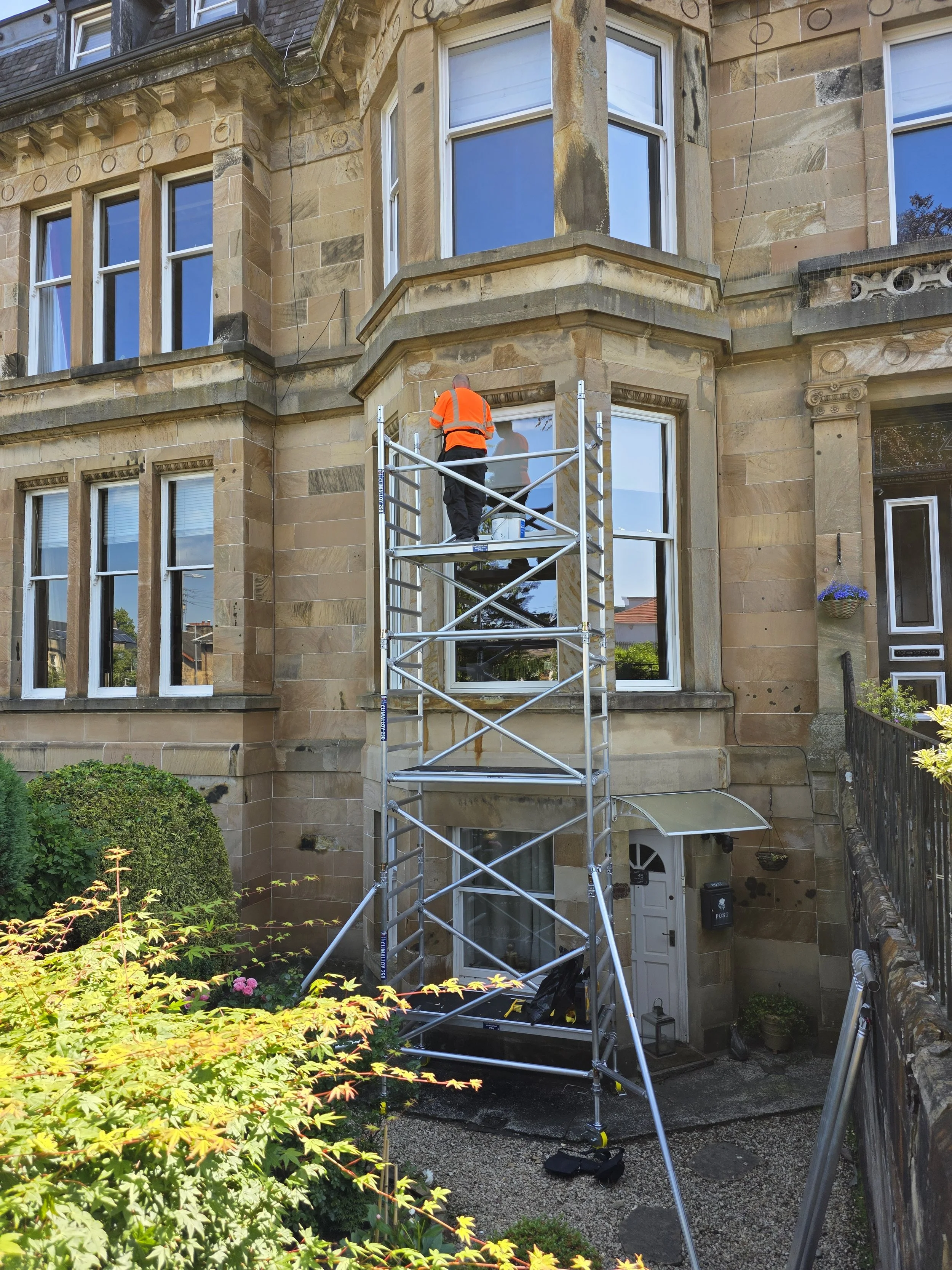 A worker in an orange safety vest and black pants is standing on a scaffold cleaning windows of a multi-story beige brick building with large bay windows and decorative architectural details.