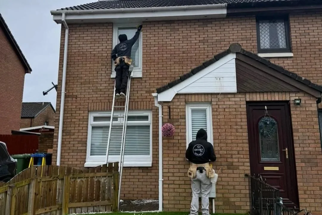Two workers in black hoodies installing a window on the upper floor of a brick house, one on a ladder and the other standing on the ground.