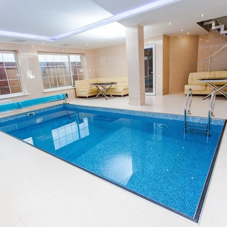 Indoor swimming pool with blue tiles, metal handrails, and beige seating areas near large windows.