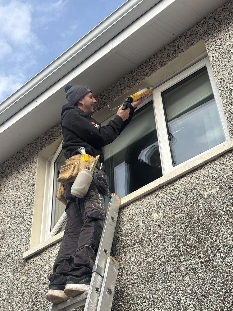 A worker standing on a ladder cleaning the exterior window of a house with a caulking gun in hand and a tool belt around his waist.