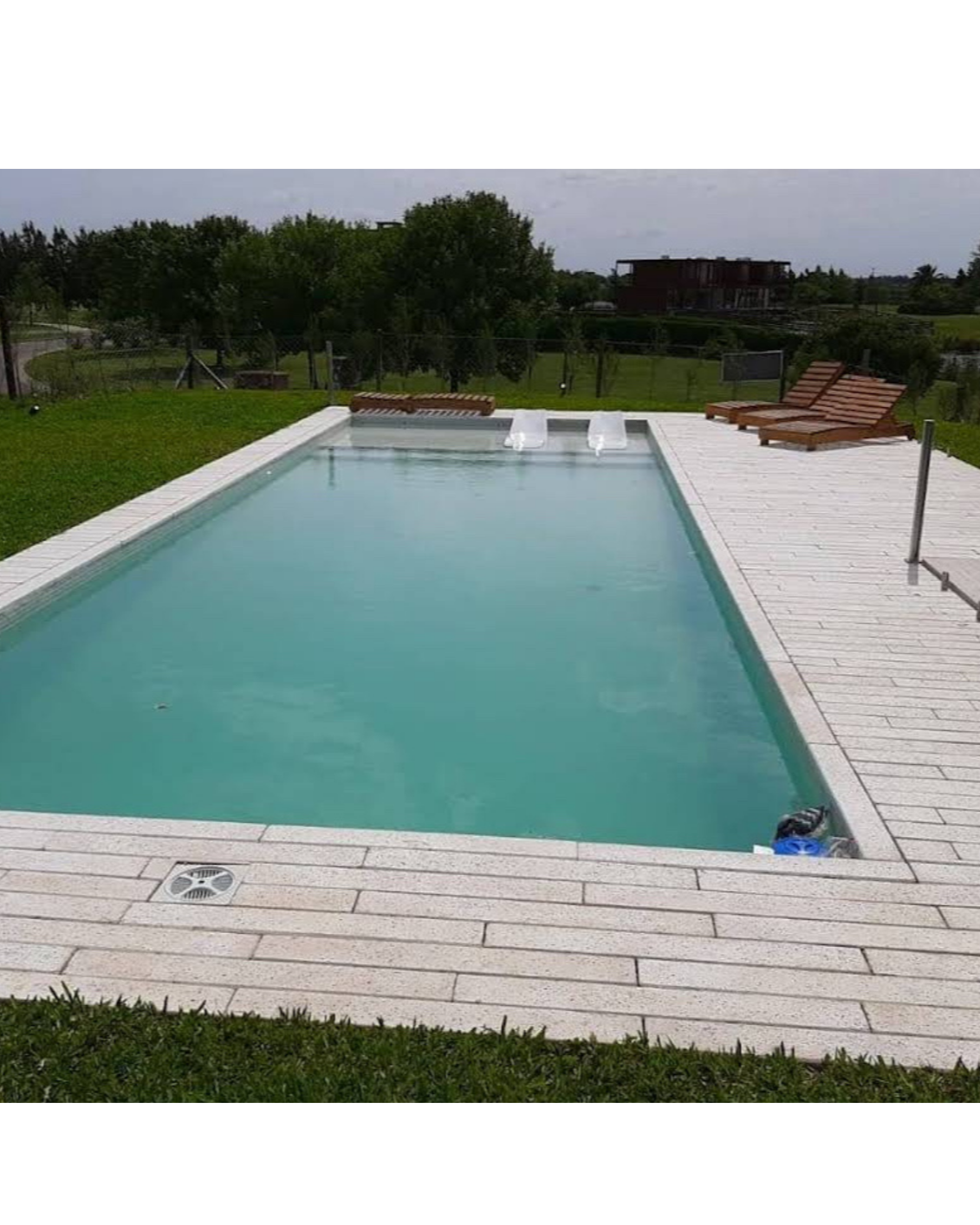 Piscina de tamaño mediano con borde de ladrillo y dos toboganes en un área al aire libre con césped y árboles, con tumbonas de madera a la derecha.
