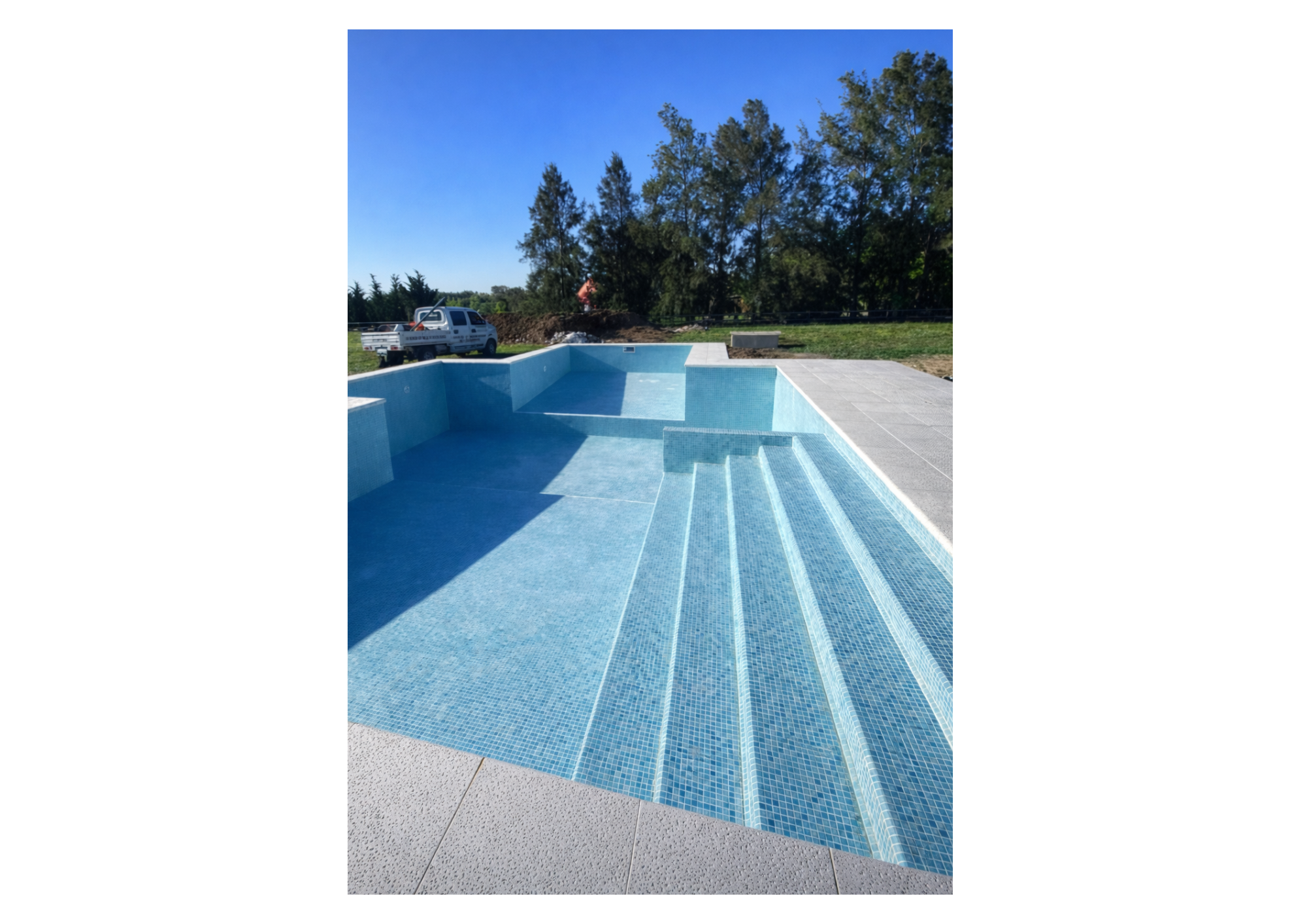 Piscina de niños con escaleras y mosaicos azules en un área al aire libre, con un fondo de árboles y un cielo despejado.