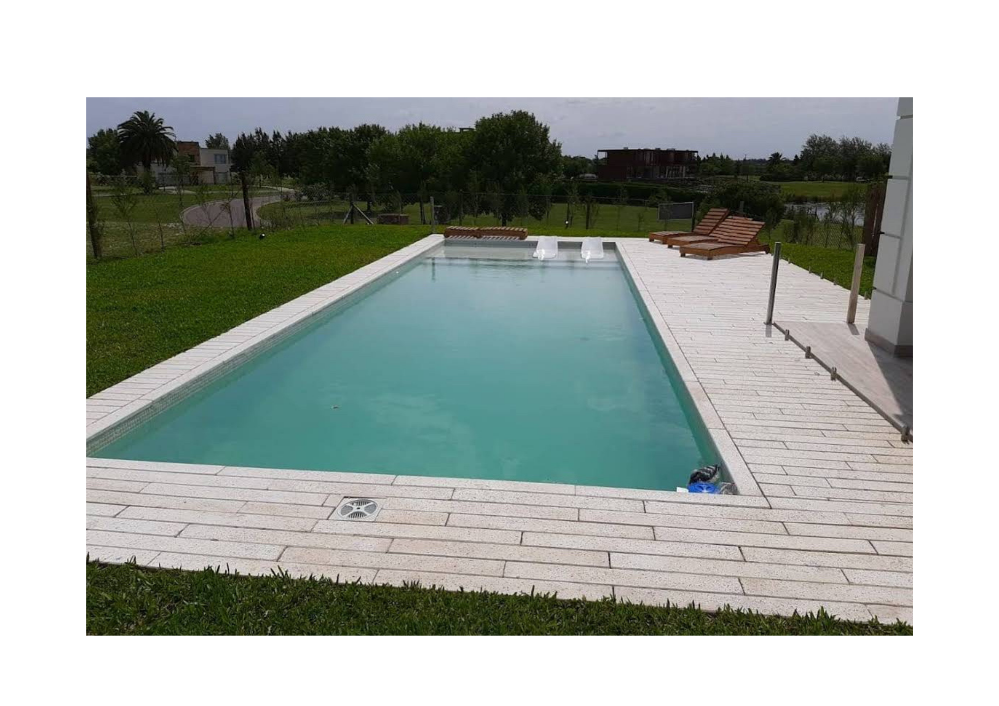 Piscina rectangular con agua clara, rodeada de piso de losas blancas, con jardín verde y árboles al fondo, y tumbonas en la esquina