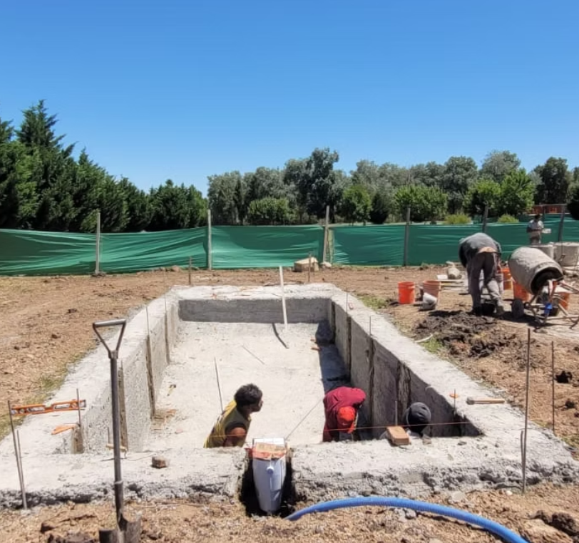 Trabajadores construyendo una piscina de cimentación de concreto en un campo con árboles y un cielo despejado.