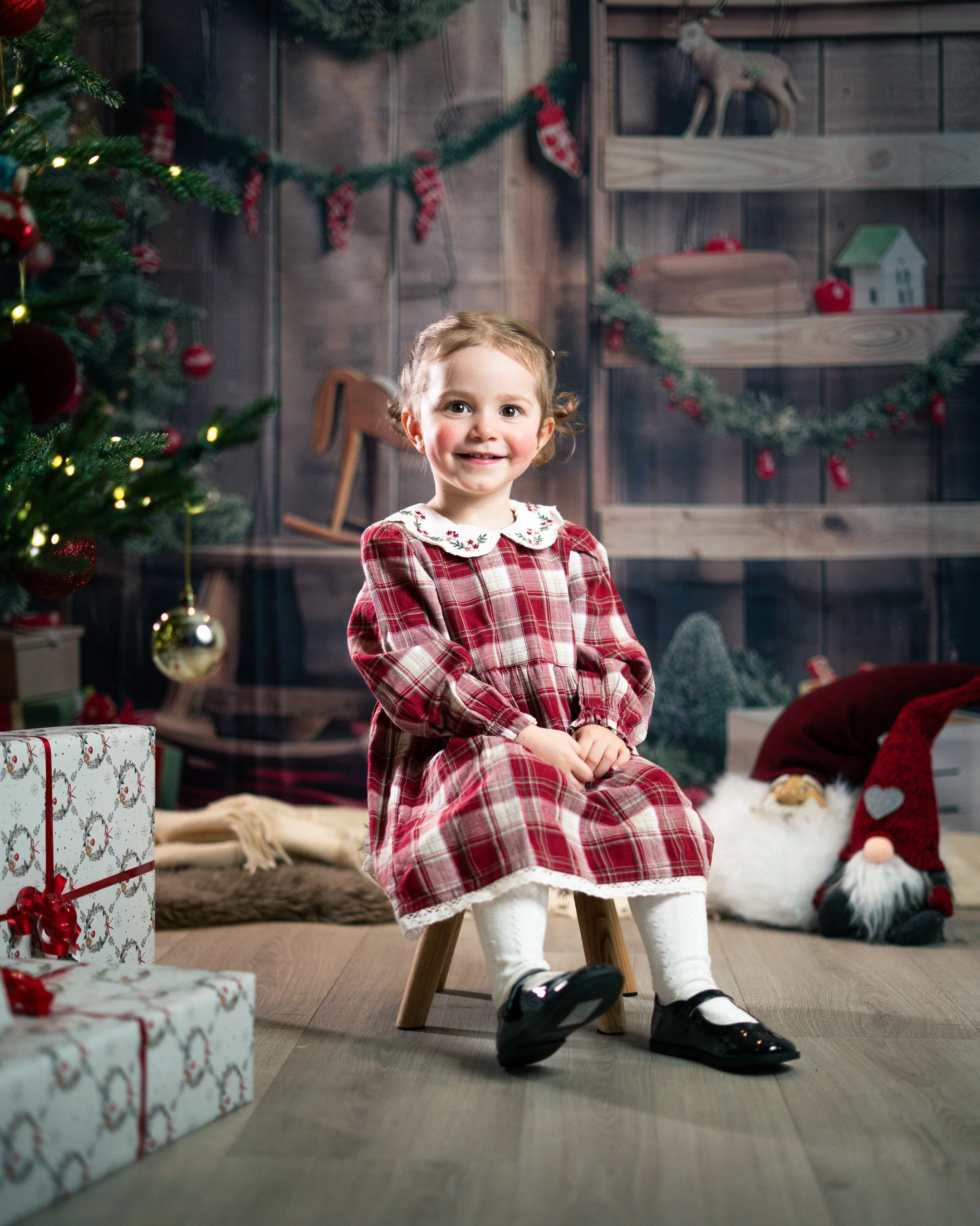 Liten jente i juleantrekk sitter på en liten benk i et julepyntet rom med juletre, gaveesker og juledekorasjoner.