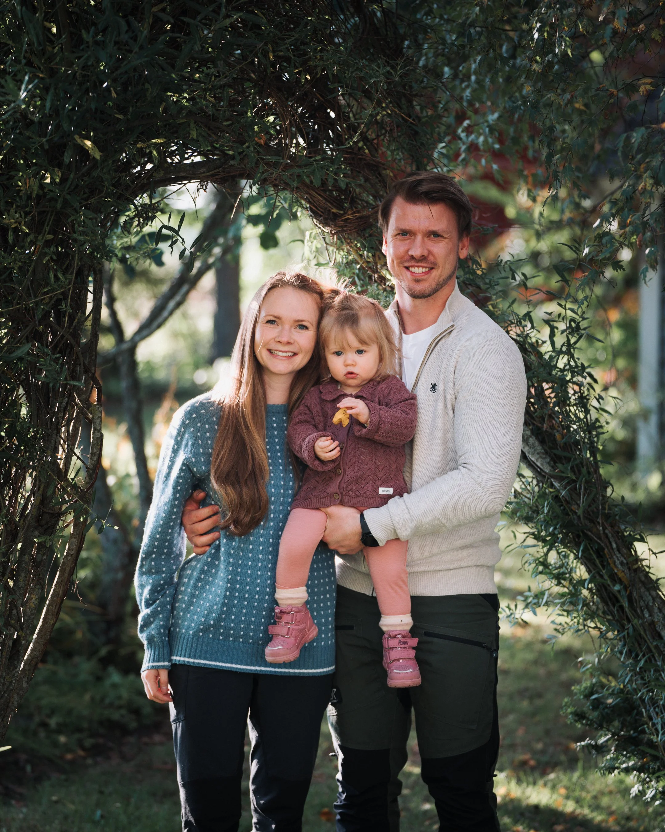 En familie med to voksne og ett barn smiler og står under en trebue i en hage med grønne trær og busker.