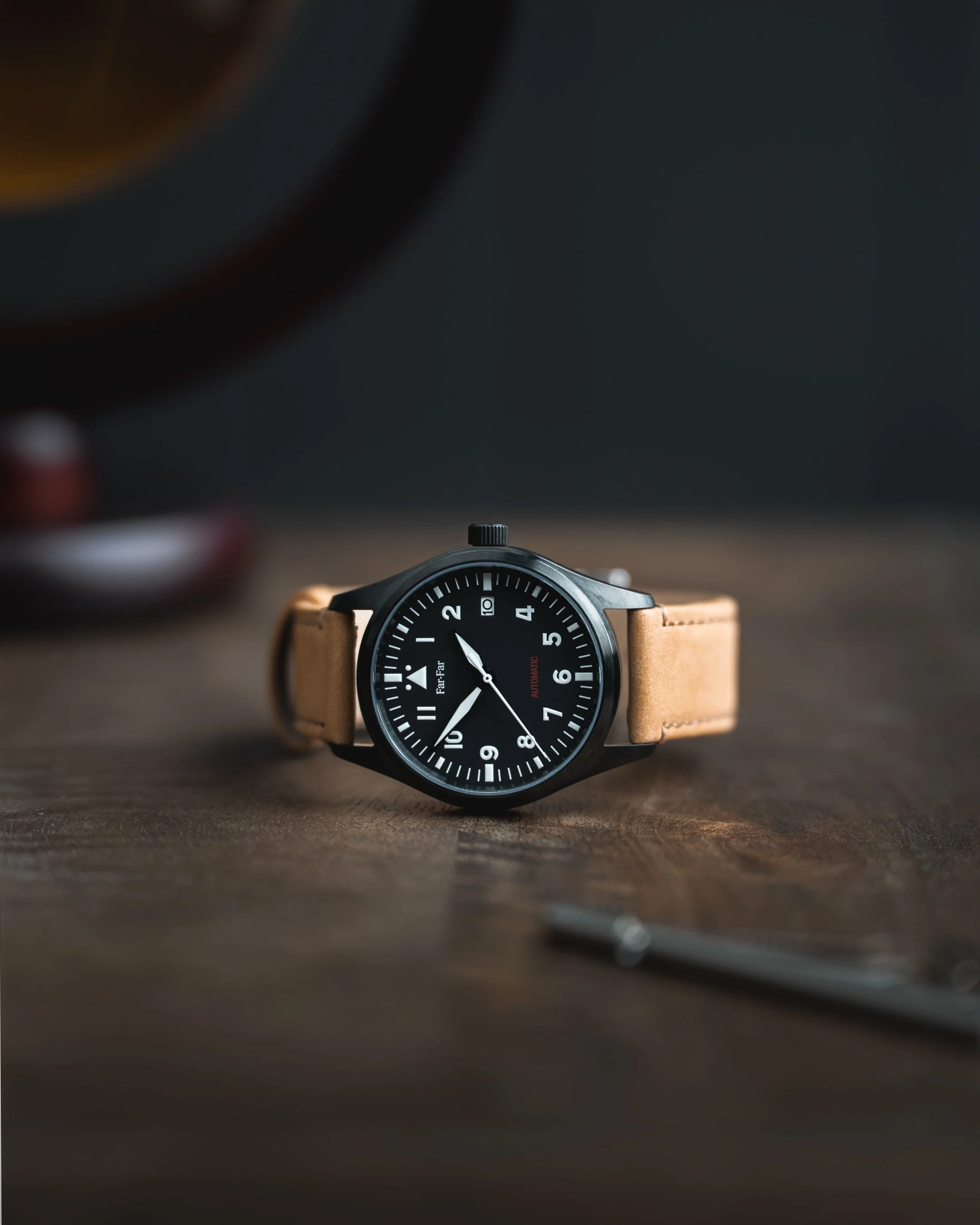 Et svart armbåndsur med brun lærrem ligger på en treoverflate. Det er en klokke med sort ansikt, hvite tall og visere, og teksten 'FattFar' på ansiktet.