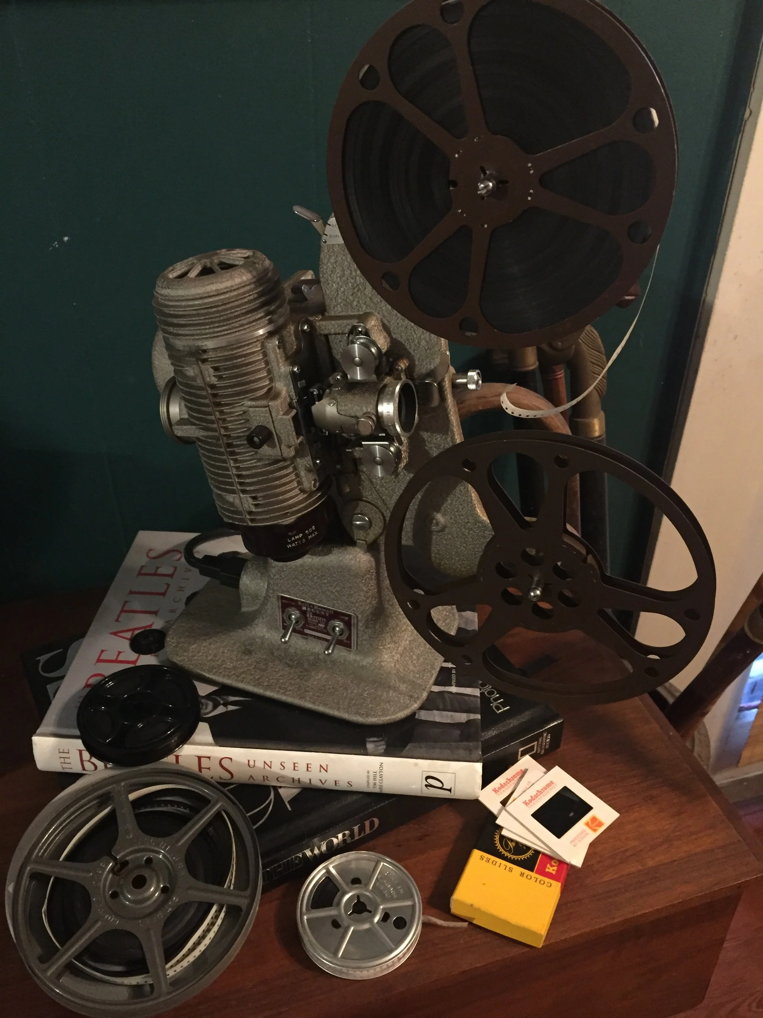 A vintage film projector with two large reels, placed on a table alongside two smaller reels, a black film reel holder, film cartridges, and books about architecture and history.