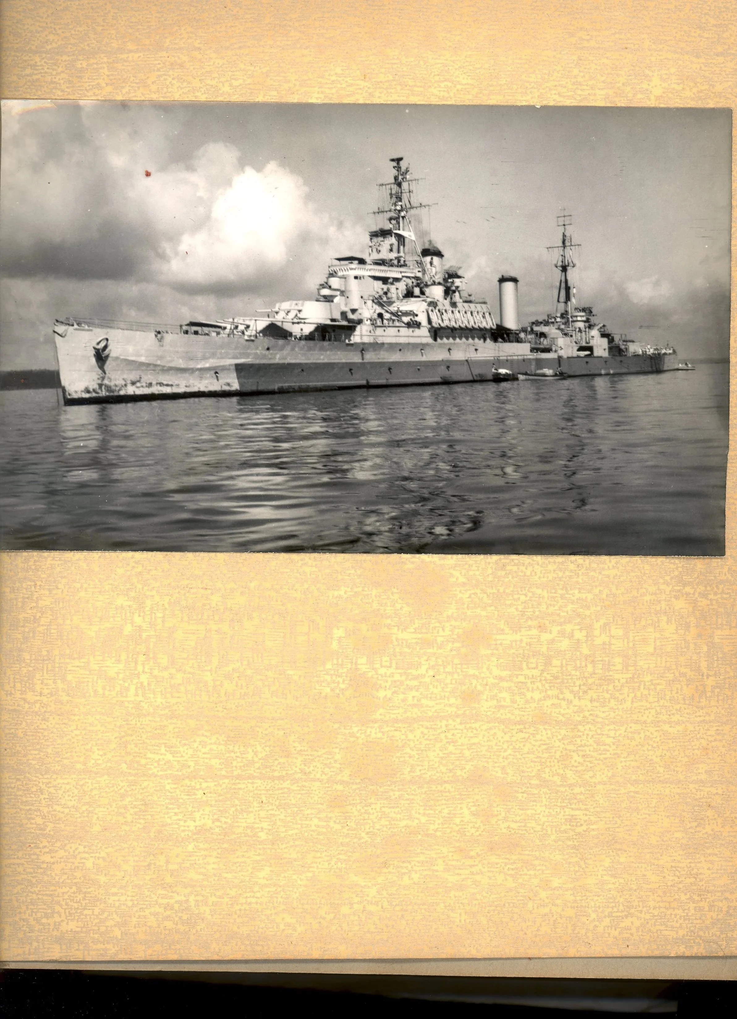 Black and white photograph of a large naval battleship on calm water, with cloudy sky in the background.