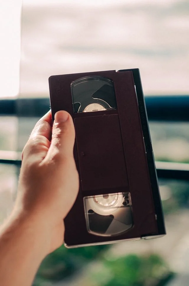 A hand holding a VHS tape indoors with a window and blurred landscape in the background.