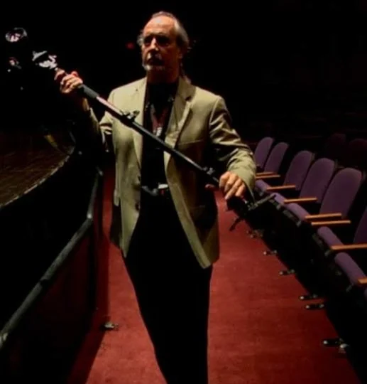 A man in a beige jacket holding a boom microphone inside an empty theater with purple seats.