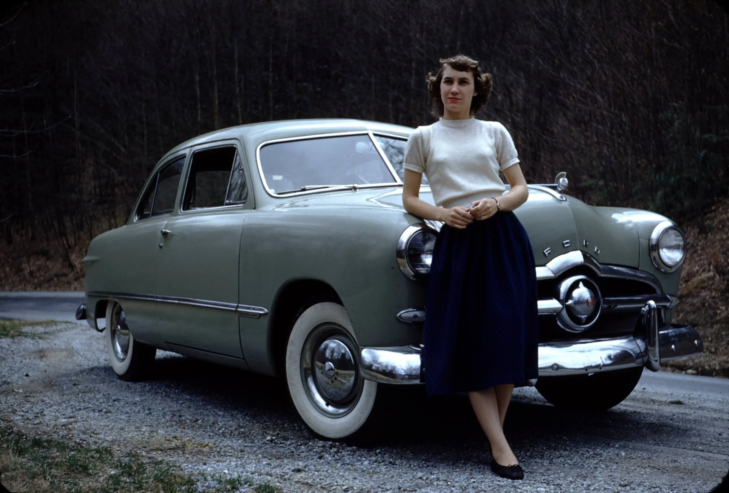 A woman with wavy brown hair standing beside a vintage green Ford car on a rural road with a wooded background.