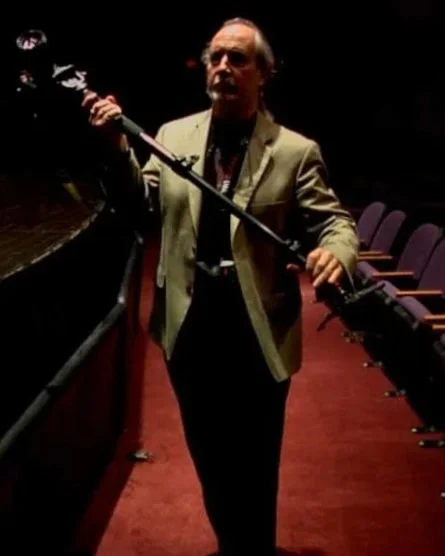 A man in a beige blazer and black shirt holding a boom microphone in a theater or auditorium.