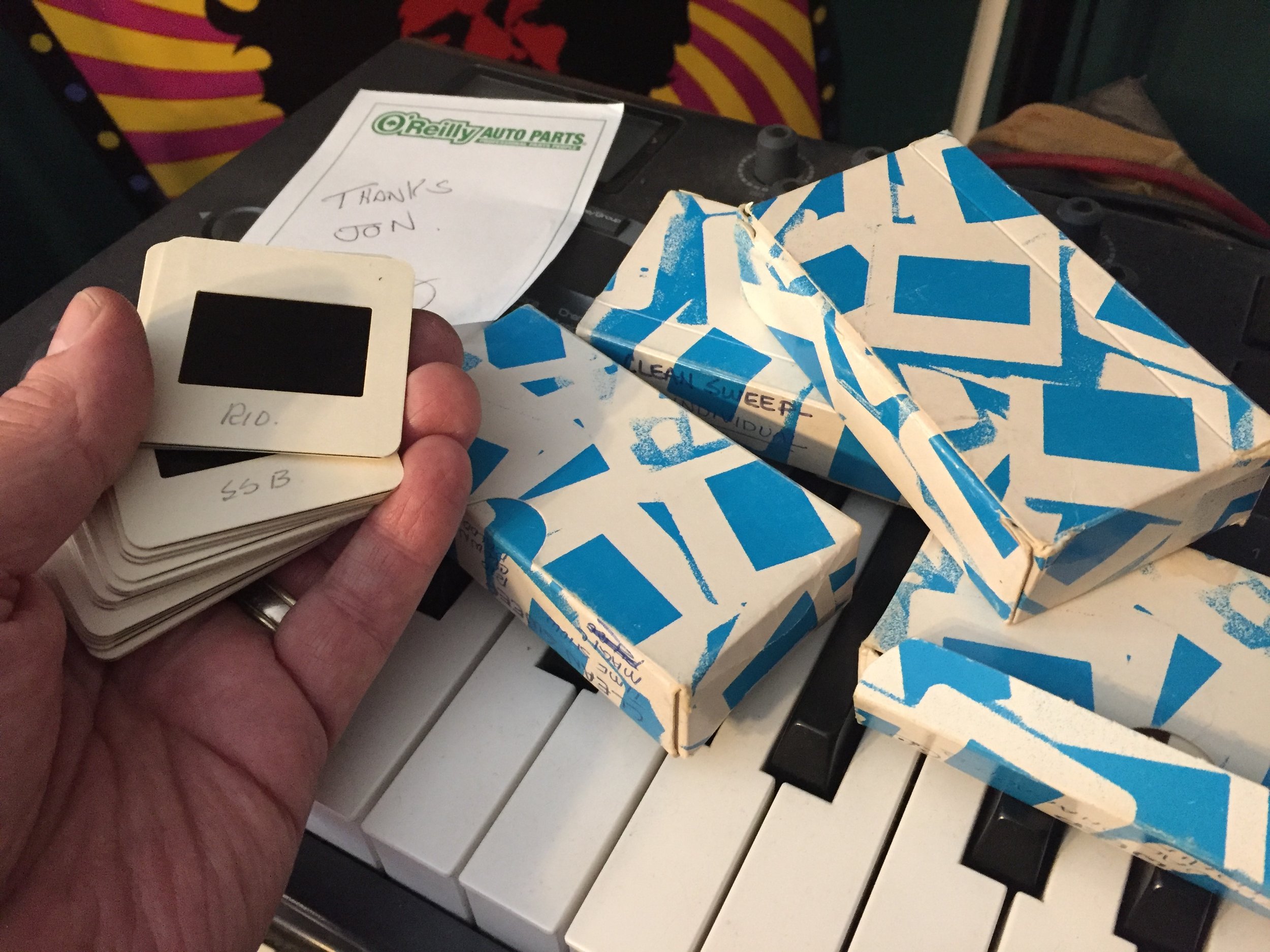 Hand holding a stack of slide mounts with handwritten labels, resting on a keyboard. Behind are several blue and white patterned boxes, a note with the O'Reilly Auto Parts logo, and some other objects.