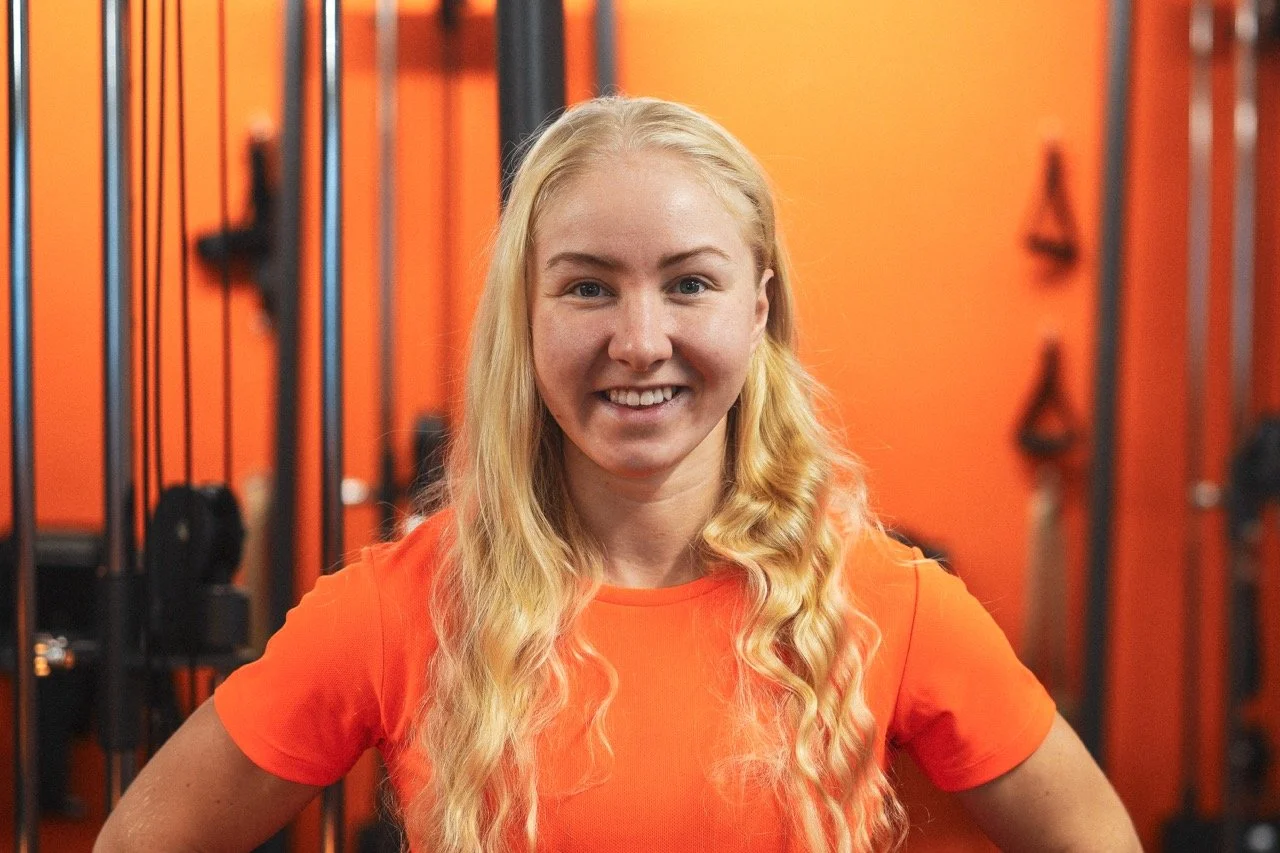 Junge Frau in einem Fitnessstudio, trägt ein oranges T-Shirt, lächelt und hat lange blonde, lockige Haare.