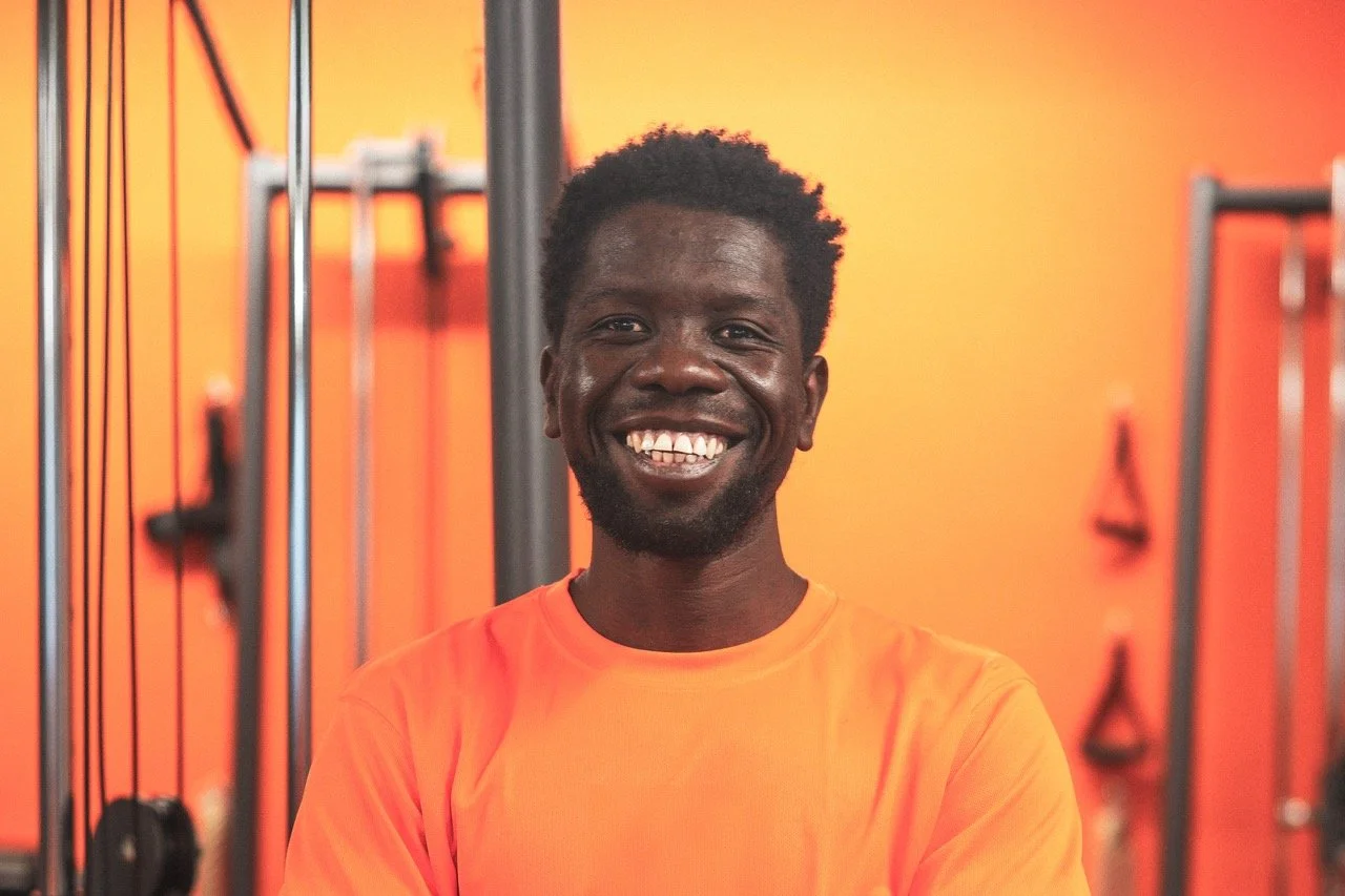 Lächelnder Mann trägt orange T-Shirt vor einem orangefarbenen Hintergrund mit Fitnessgeräten.