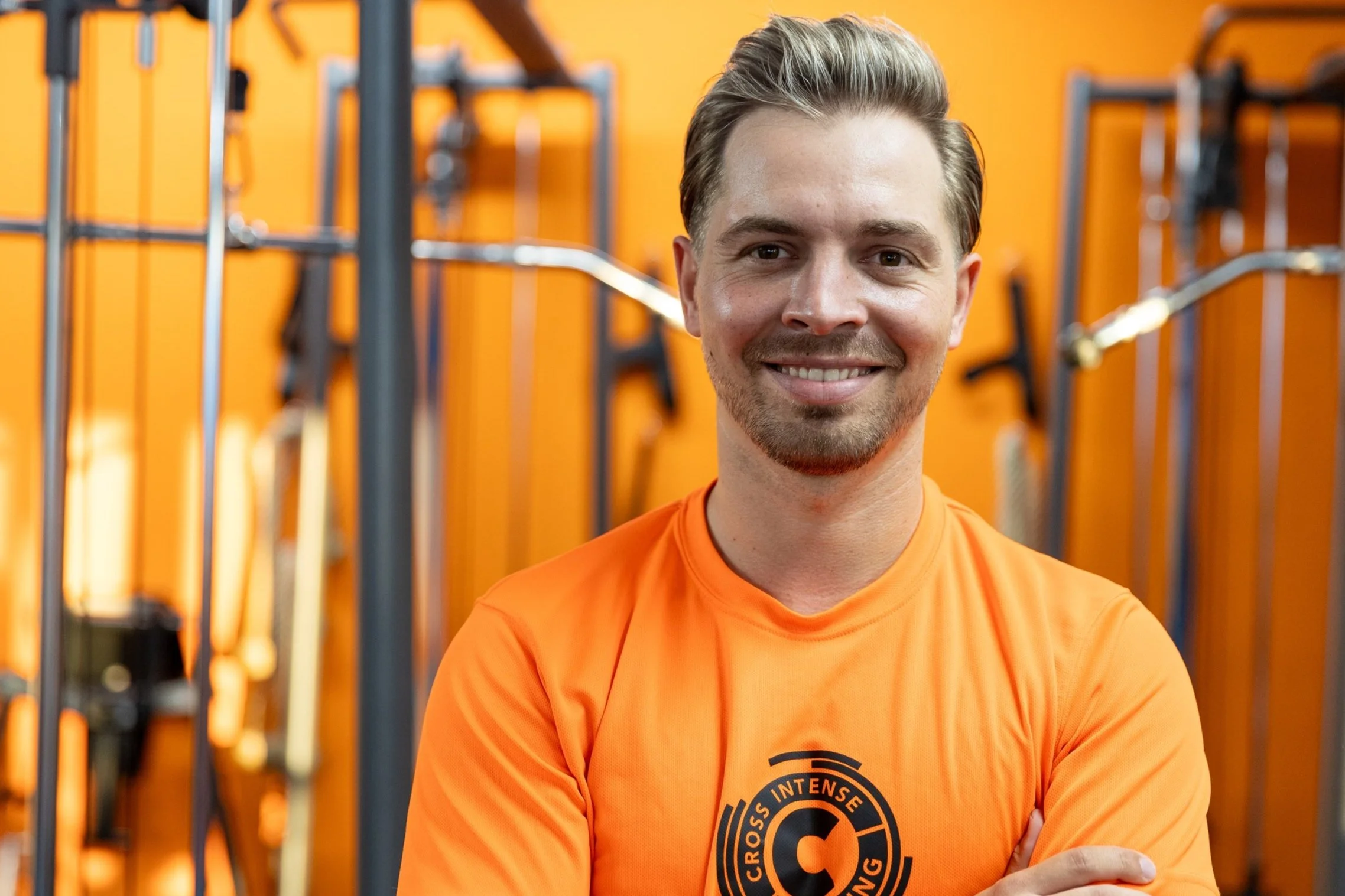 Mann trägt orange T-Shirt mit Cross Fit Logo, lächelt, steht in einem Fitnessstudio mit orangefarbenen Wänden und Fitnessgeräten im Hintergrund.