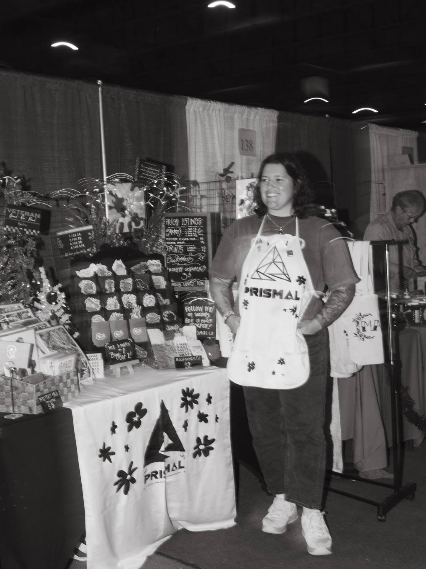 A woman standing behind a table at a craft fair, smiling and wearing an apron with a geometric design and the word "Prismal." The table displays flowers, signs, and various small items for sale, with a sign in the background listing prices.