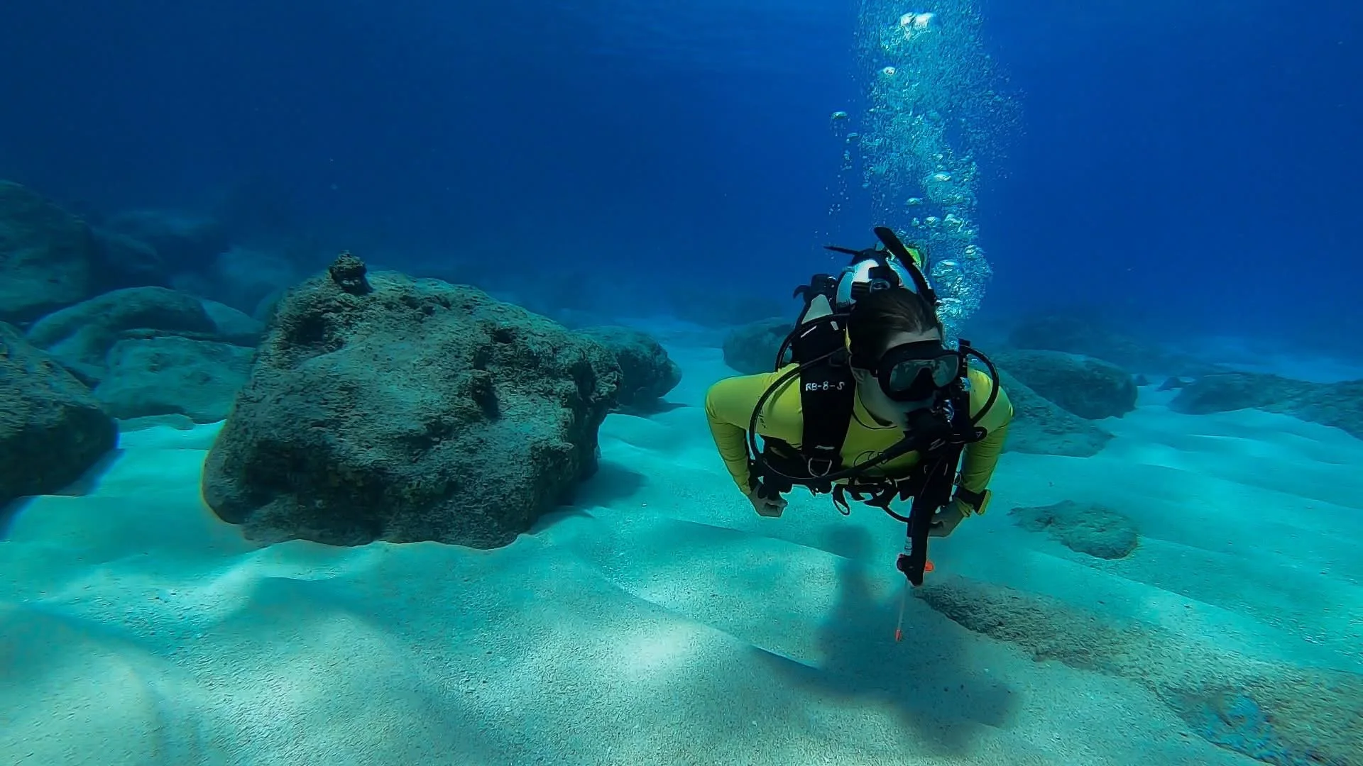 A scuba diver swimming underwater near rocks and sand with bubbles rising to the surface.