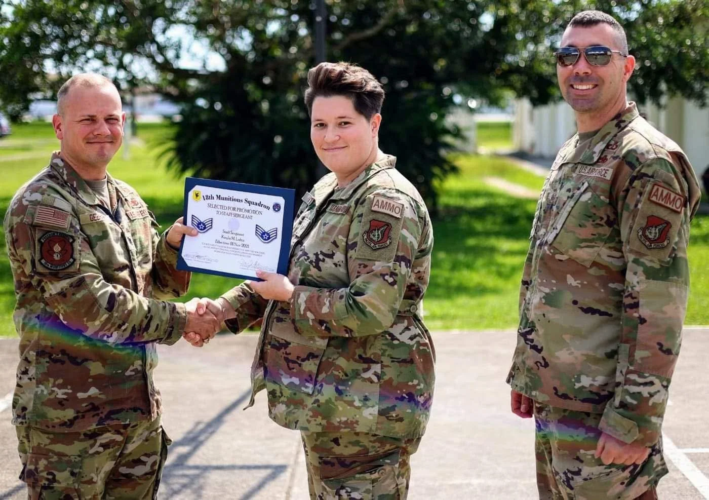 A woman in military uniform receives a promotion certificate from a man in military uniform, shaking hands, with another man in military uniform standing nearby, outdoors with trees and grass in the background.
