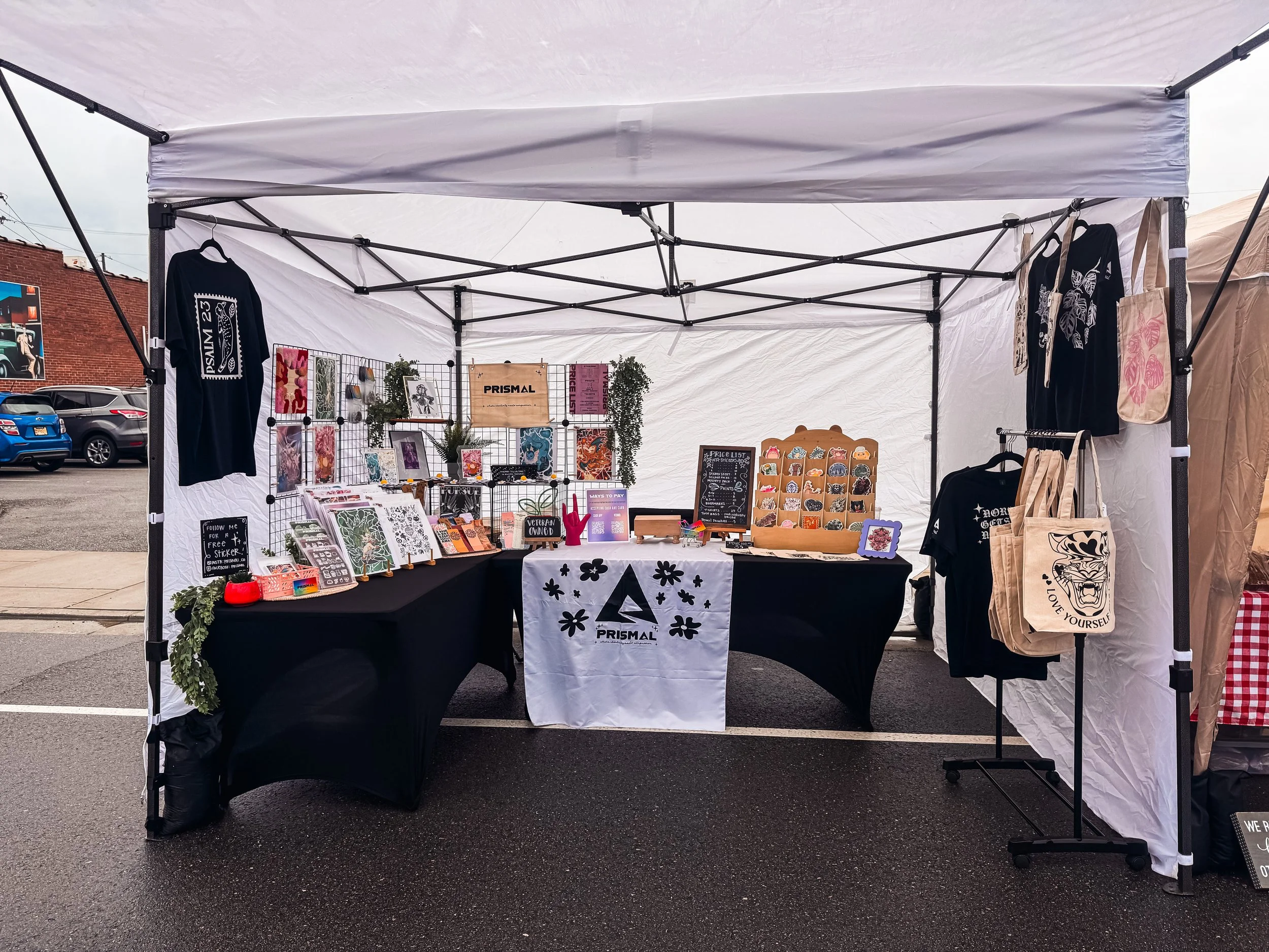 Market stall with various artwork, plants, and tote bags under a white tent, set up outdoors on a wet parking lot.