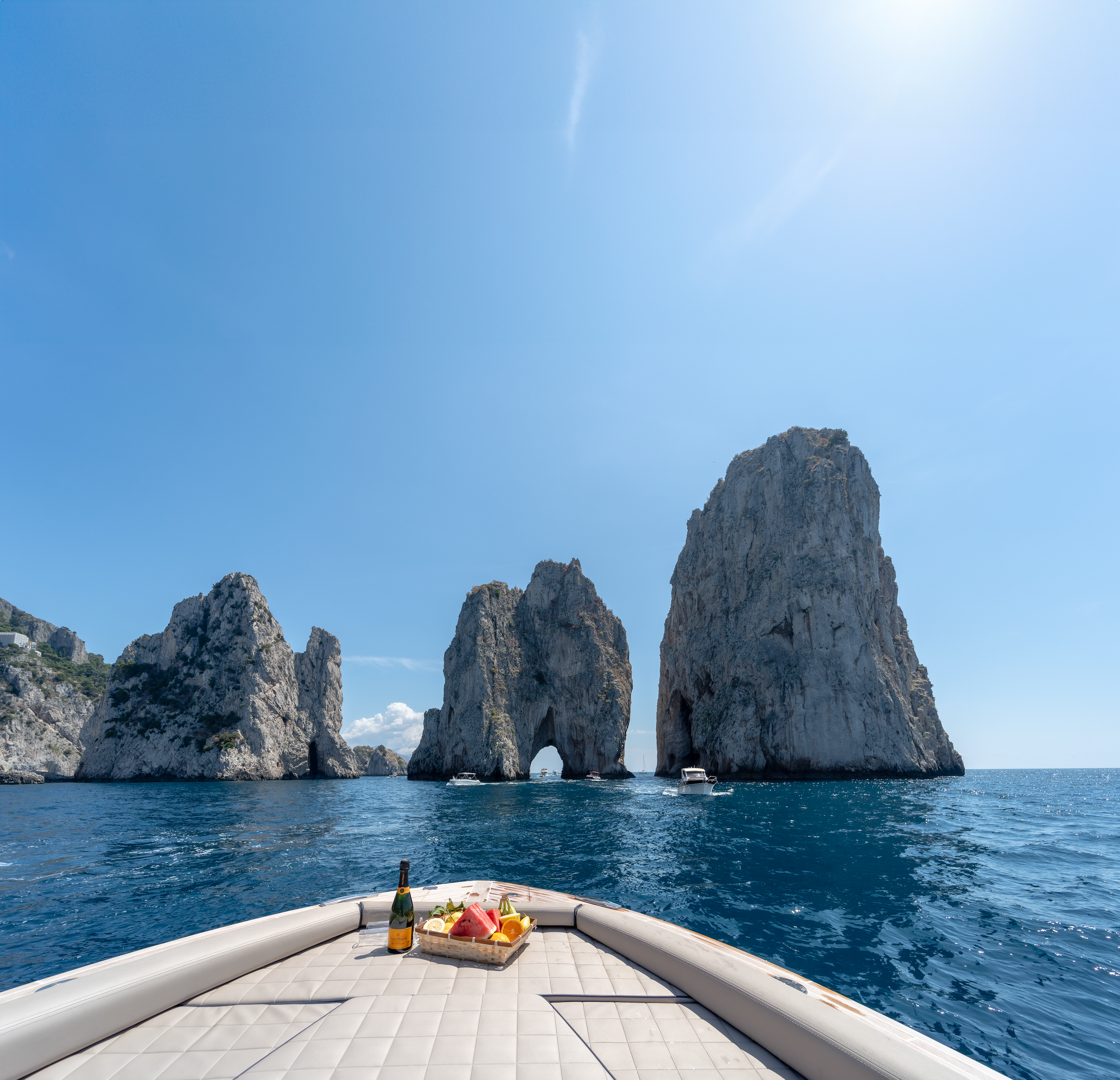 View from a boat showing three large sea stacks with a natural arch in the middle, surrounded by blue ocean and a clear blue sky.