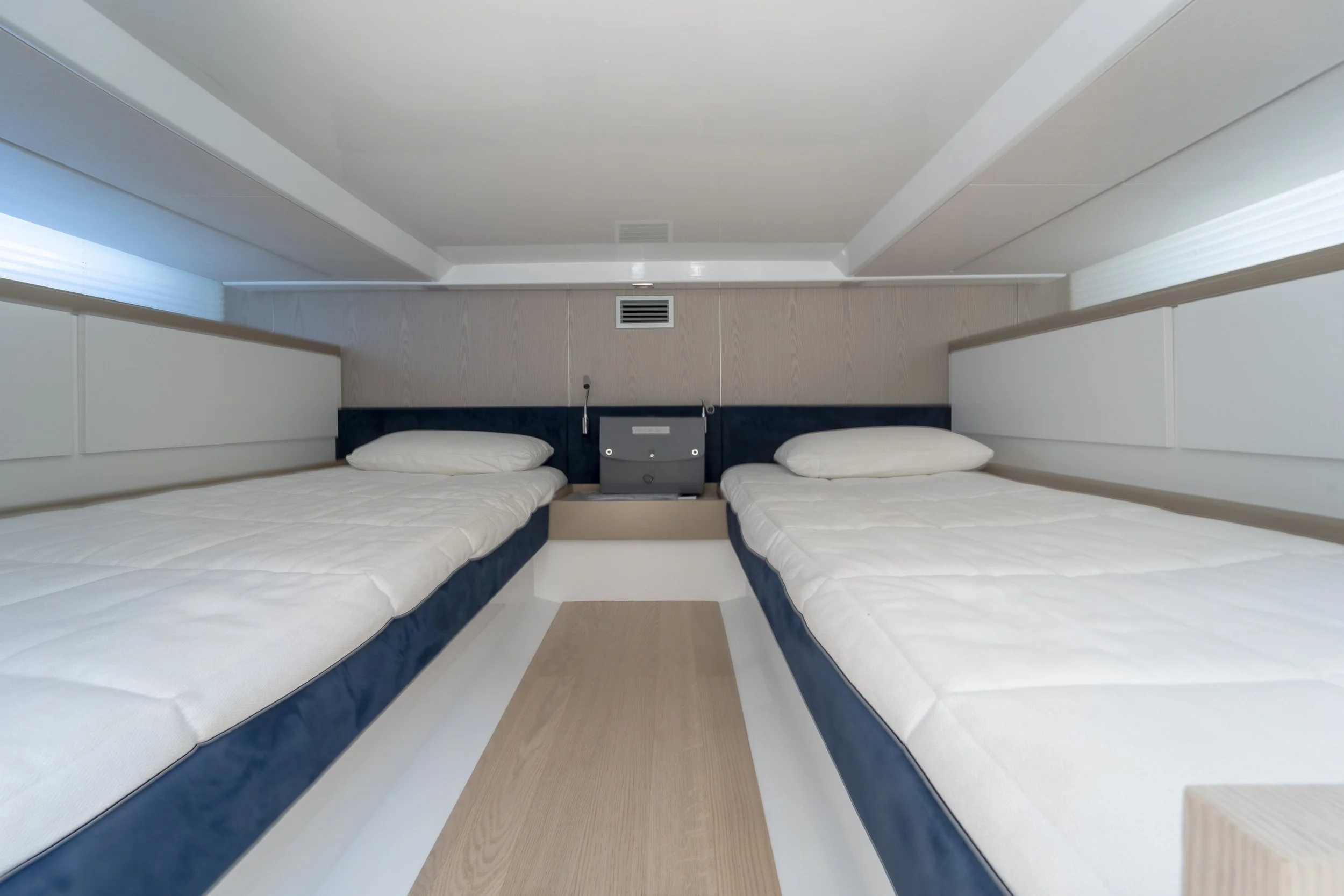 Two single beds with white linens and pillows in a minimalist hotel room, separated by a small central nightstand with a TV and electrical outlets, under a sloped ceiling with ventilation.