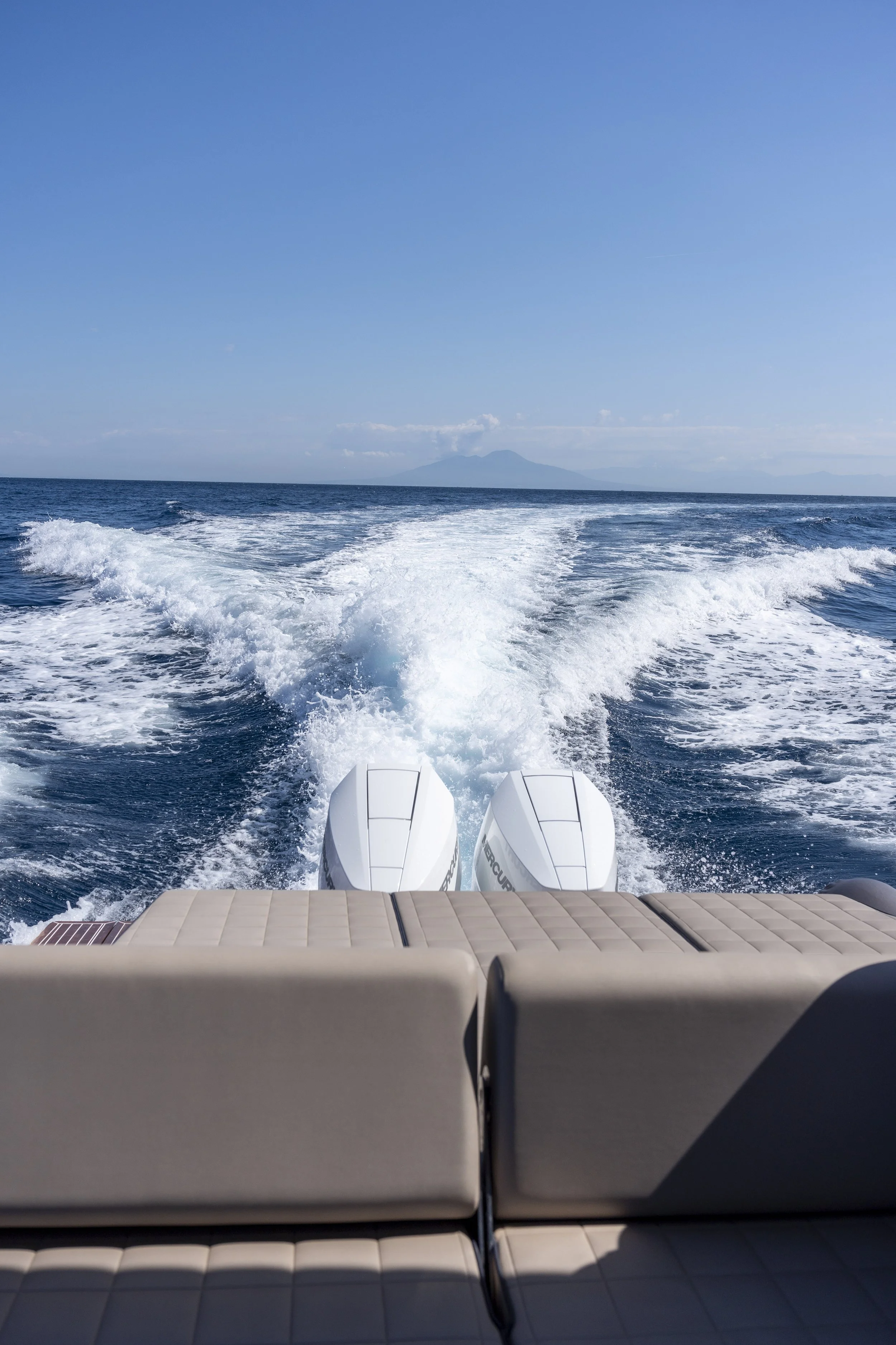 View from the back of a boat showing two outboard motors, with the boat traveling through the ocean, leaving a trail of white foam behind. A distant landmass is visible on the horizon under a clear blue sky.