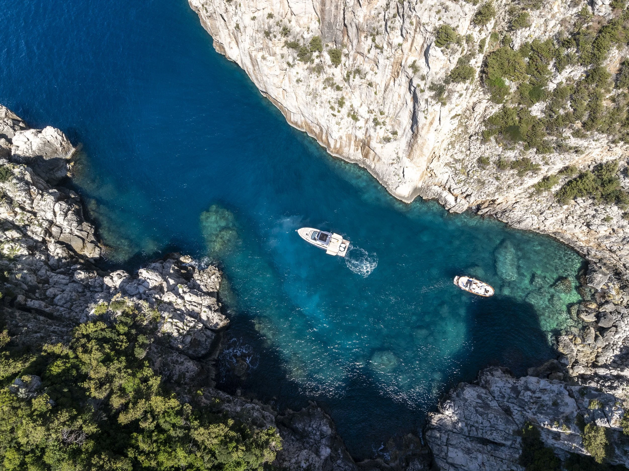 Aerial view of a boat and a small motorboat in a narrow, rocky coastal inlet with clear blue water, surrounded by steep, rocky cliffs and greenery.