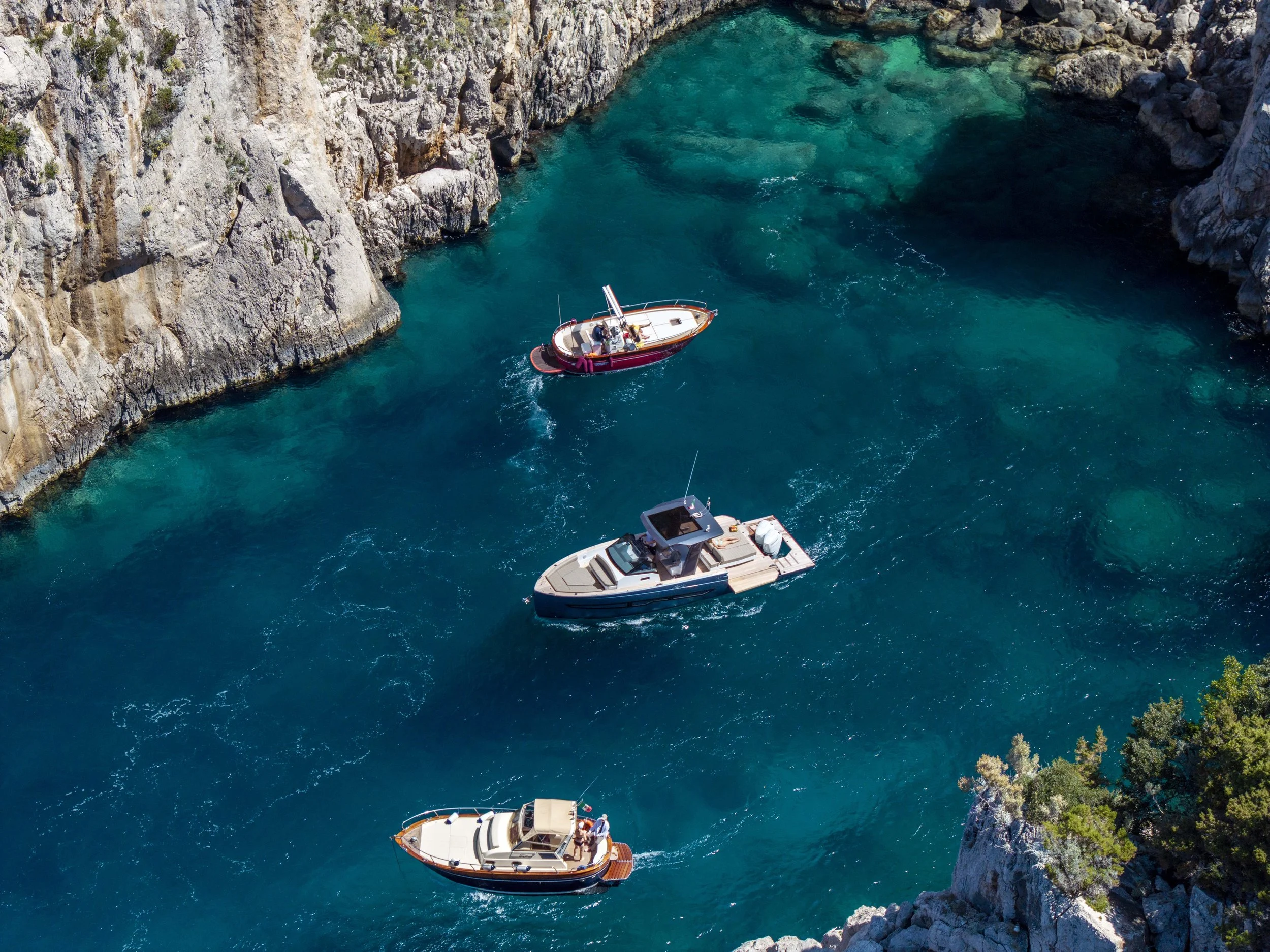 Aerial view of three boats sailing in a narrow, rocky cove with turquoise water.