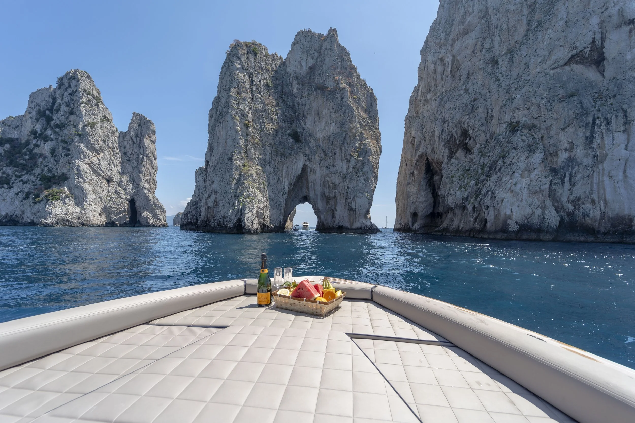 View from a boat with a padded deck, a tray of fruit and drinks, and a bottle of sparkling wine, facing large rock formations with natural arches in the water under a clear blue sky.