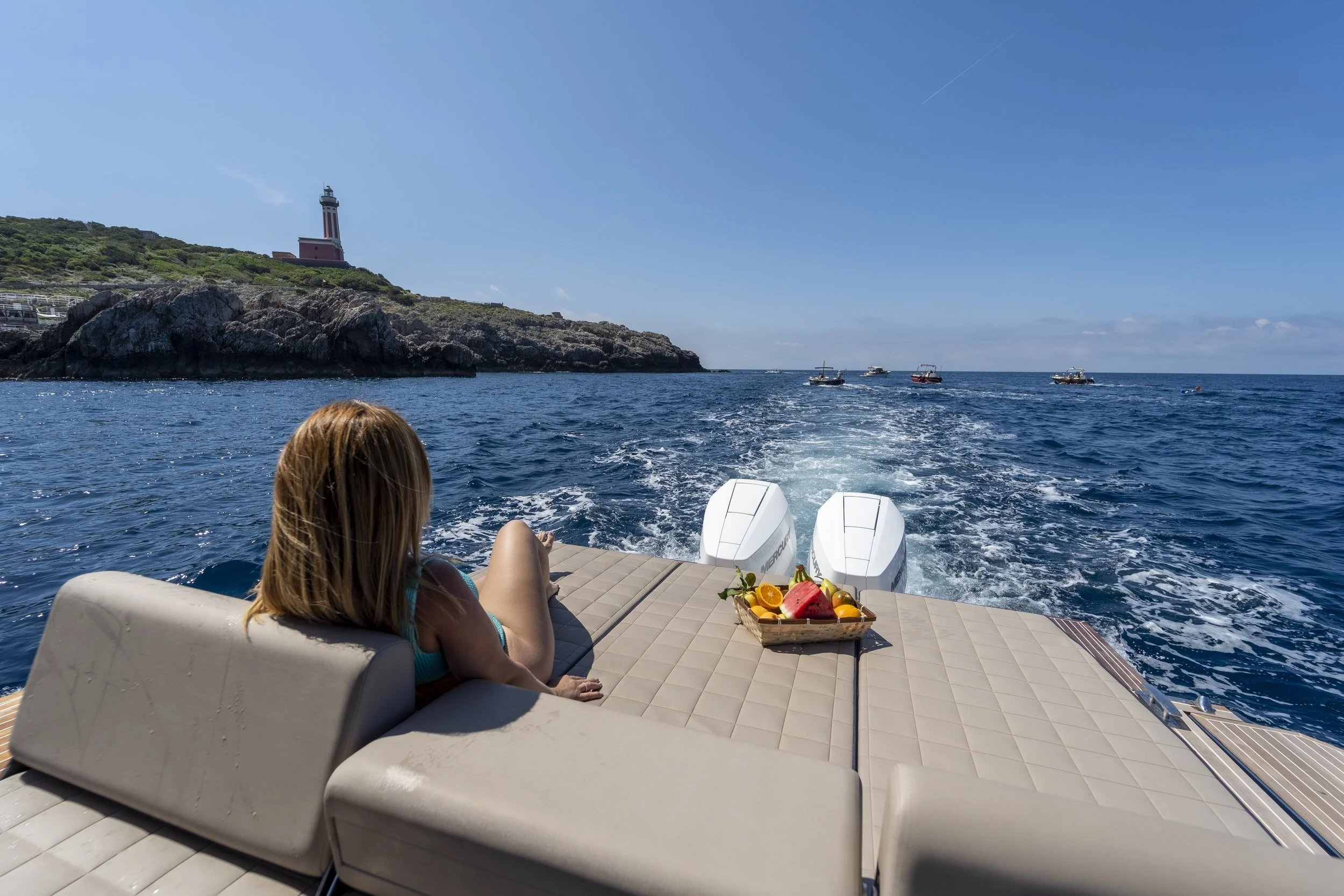 Girl relaxing on a boat with outboard motors, overlooking the ocean with a rocky coastline and lighthouse in the distance, and a basket of fruit on the deck.