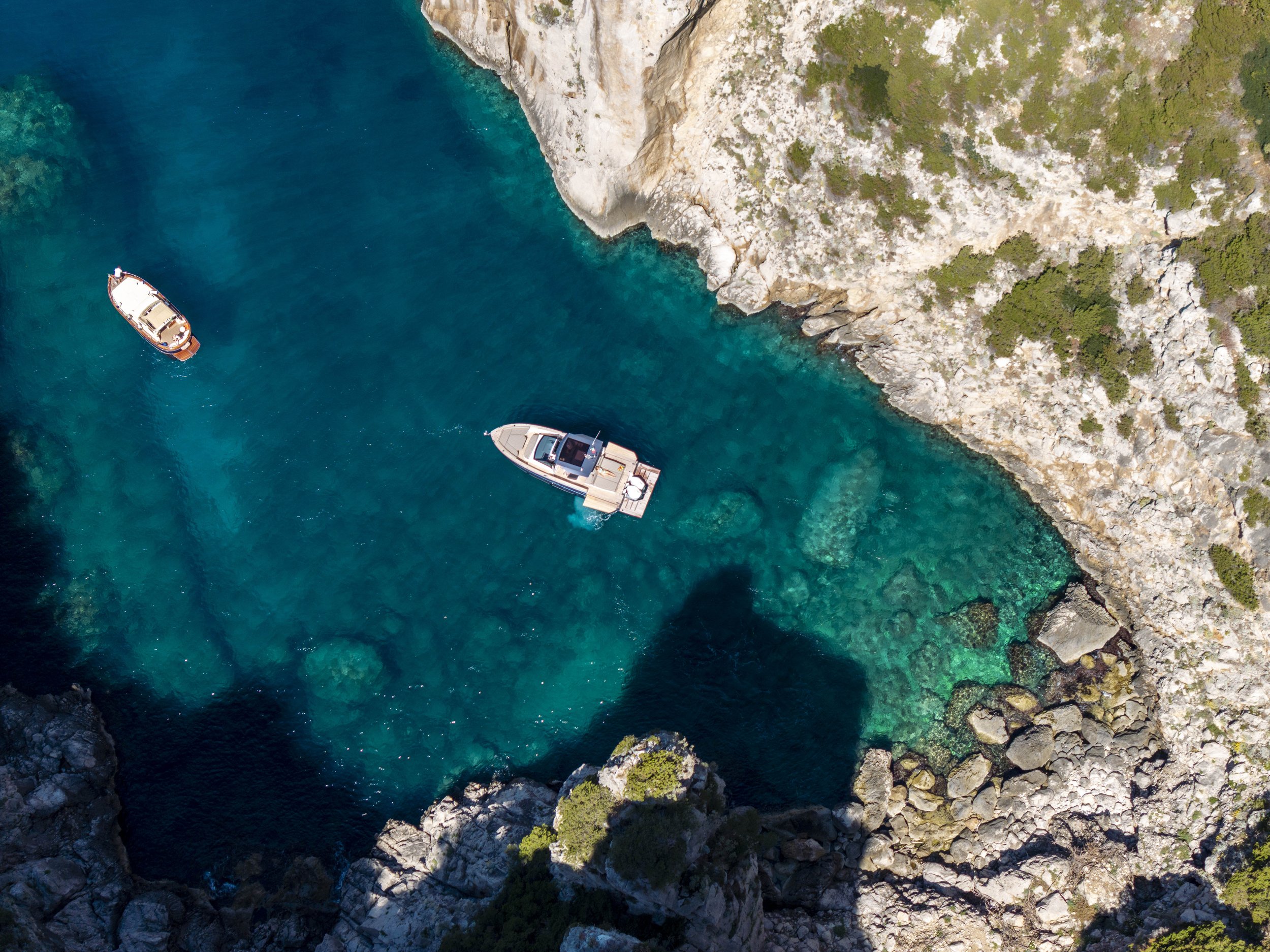 Aerial view of three boats floating in a turquoise cove surrounded by rocky cliffs with sparse vegetation.