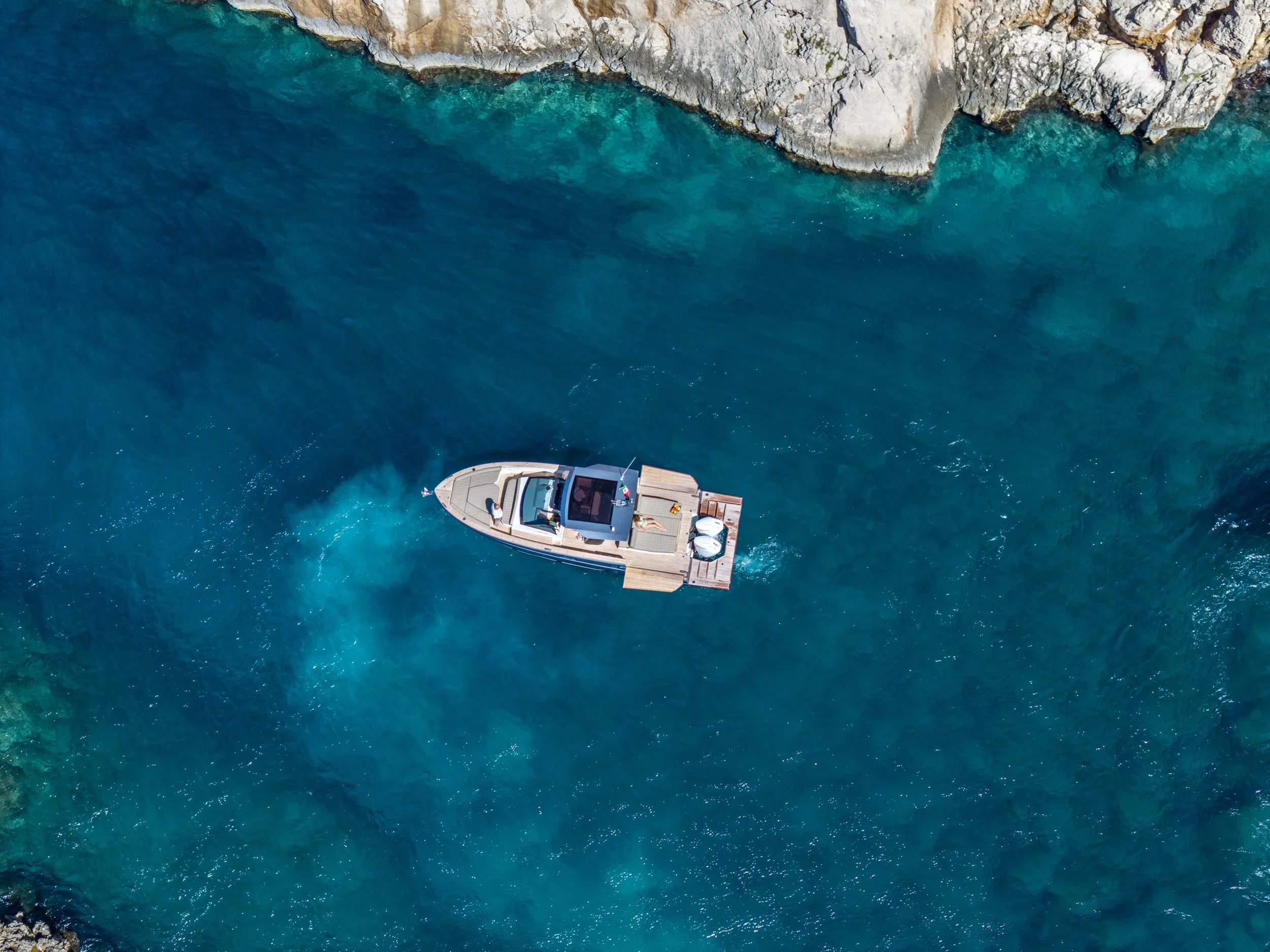 An aerial view of a boat moving through clear blue water near rocky cliffs.