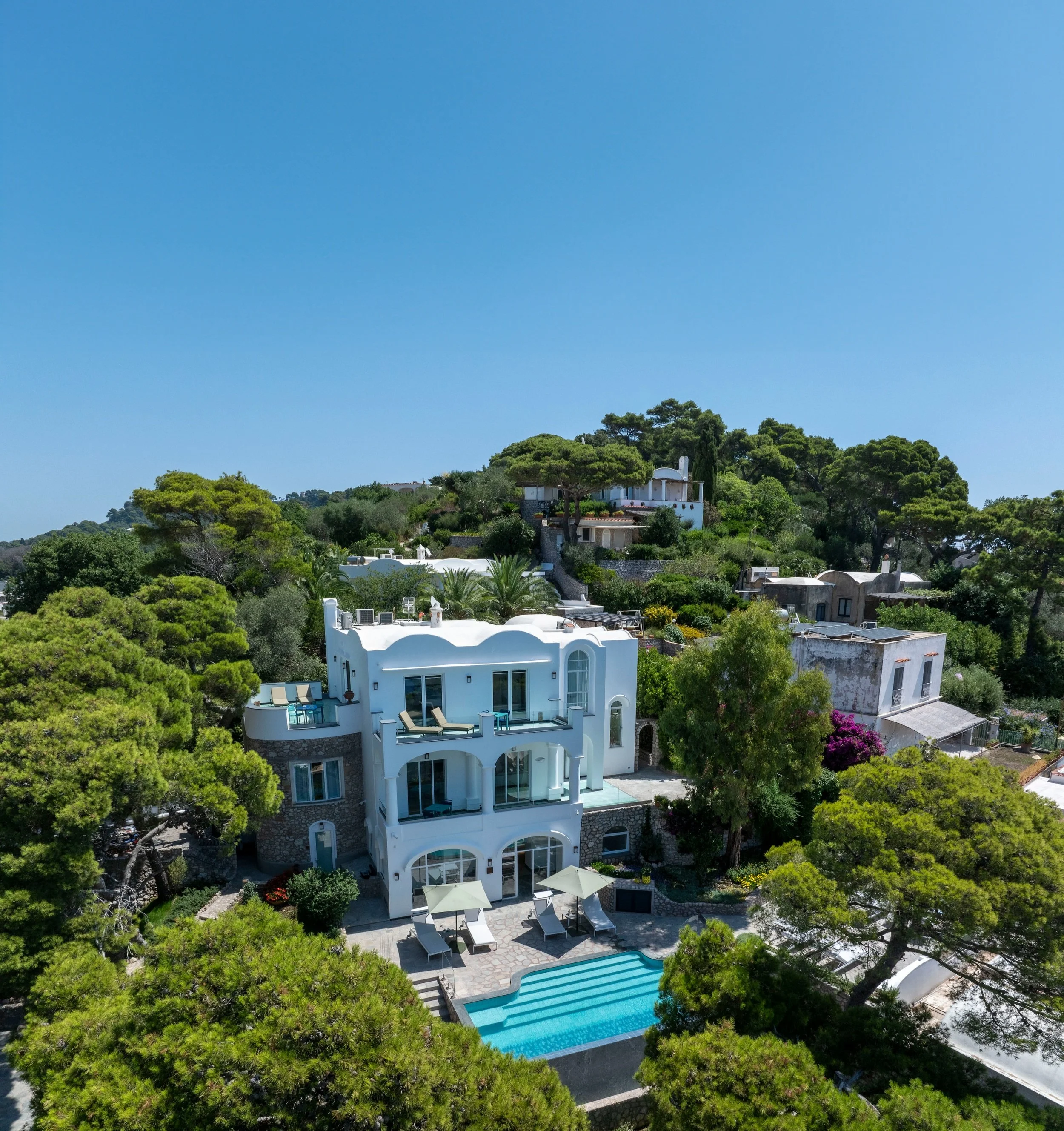 Aerial view of a luxurious white villa with multiple terraces, a swimming pool, surrounded by lush green trees, on a hill under a clear blue sky.